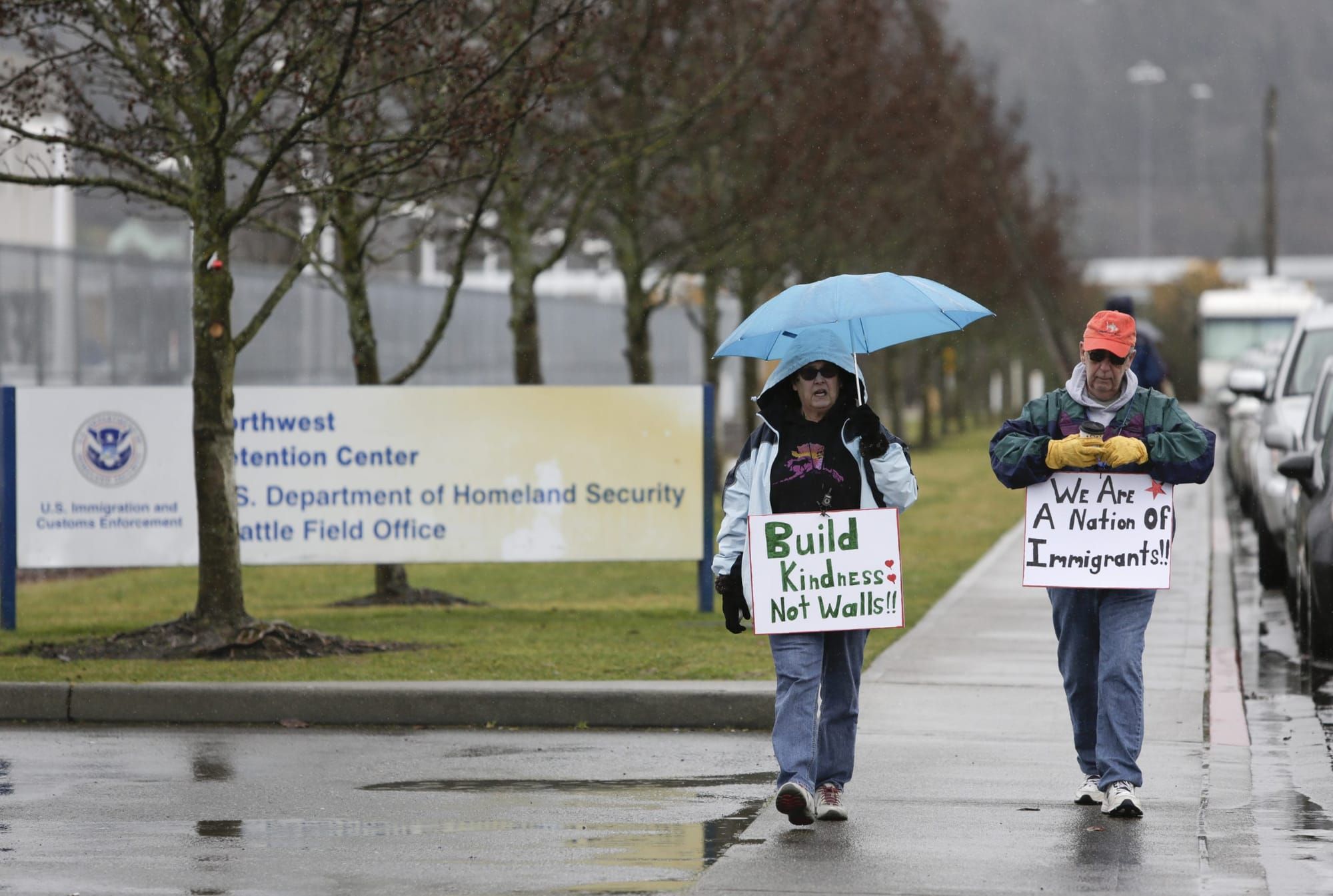 two people walk on a sidewalk past a sign for Northwest Detention Center while holding signs reading "Build kindness not wall