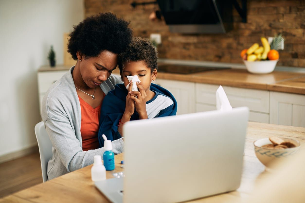 Sick African American boy blowing nose while sitting wrapped in a blanket on mother's lap at home.