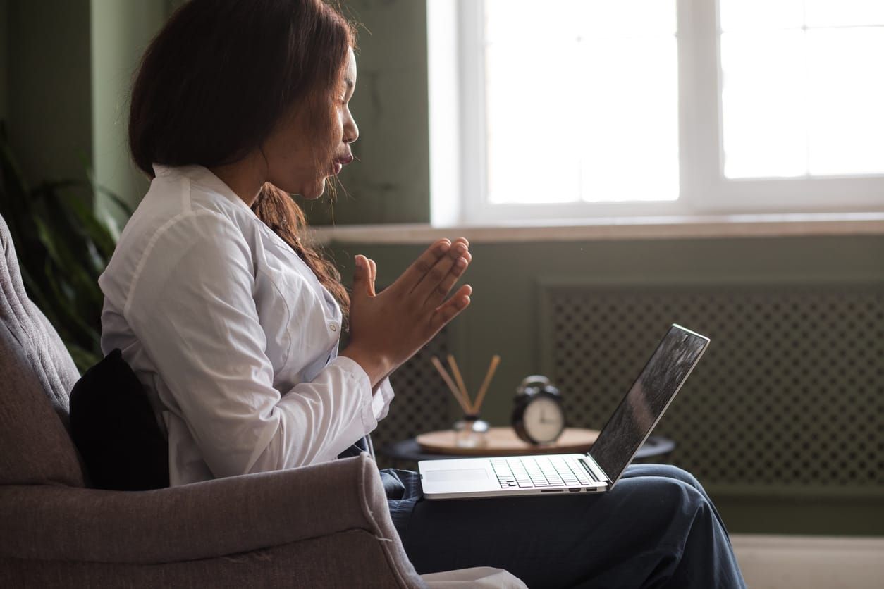 a Black femme doctor in a white long-sleeved jacket sits with a laptop in her lap, gesturing towards the screen while she tal