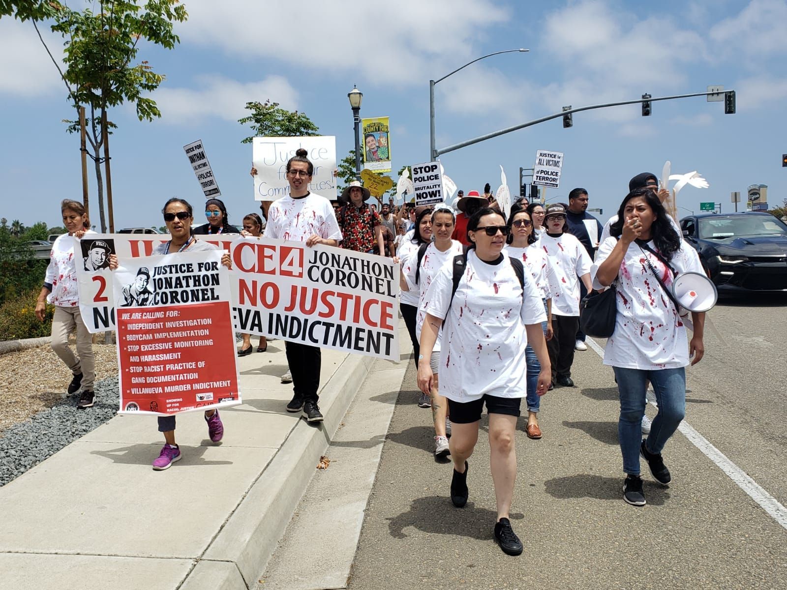 people in white T-shirts covered in decorative bloody bullet holes march down a street holding signs reading "justice for Jon