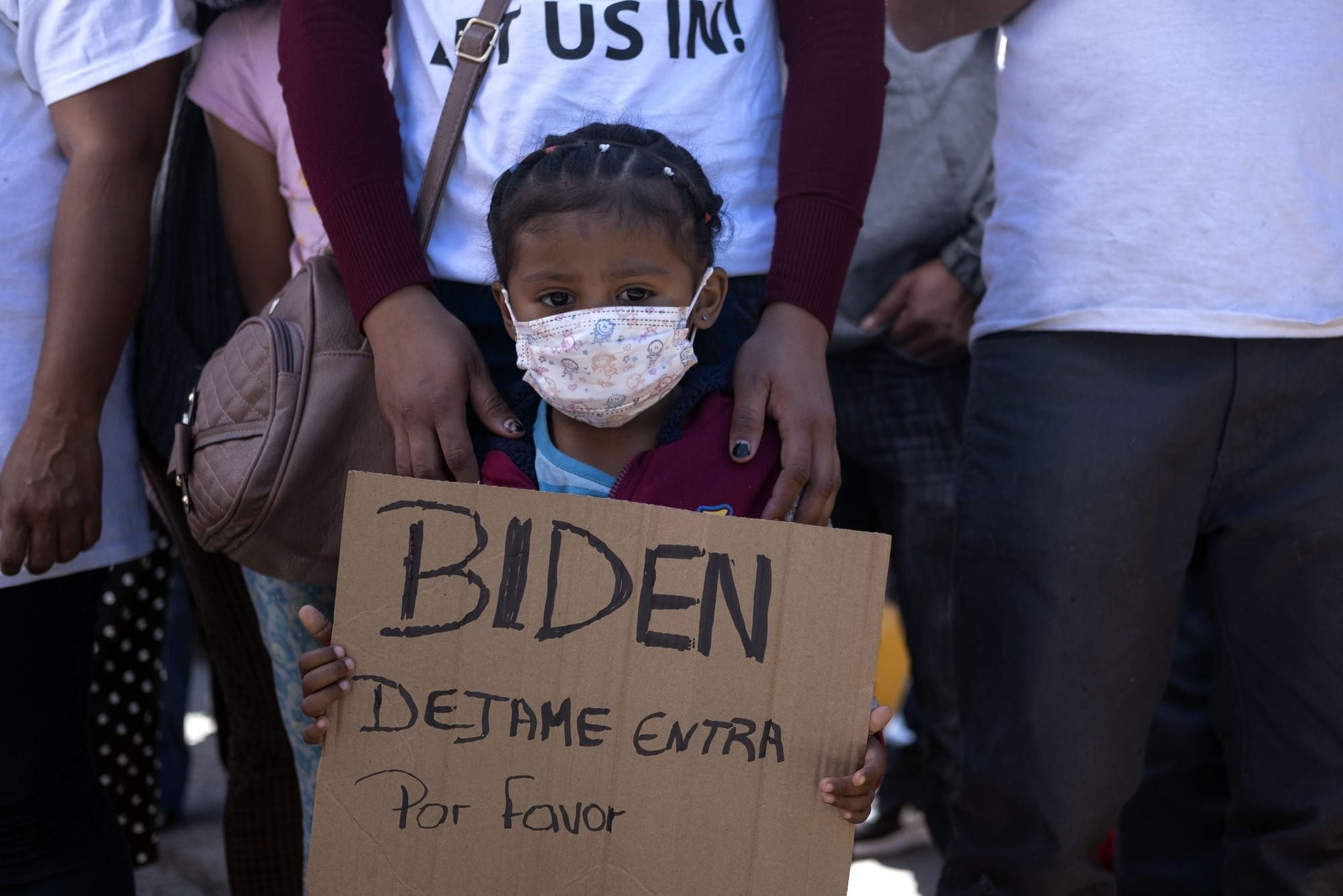 a young Honduran child holds a cardboard sign with the text "Biden dejame entra por favor"