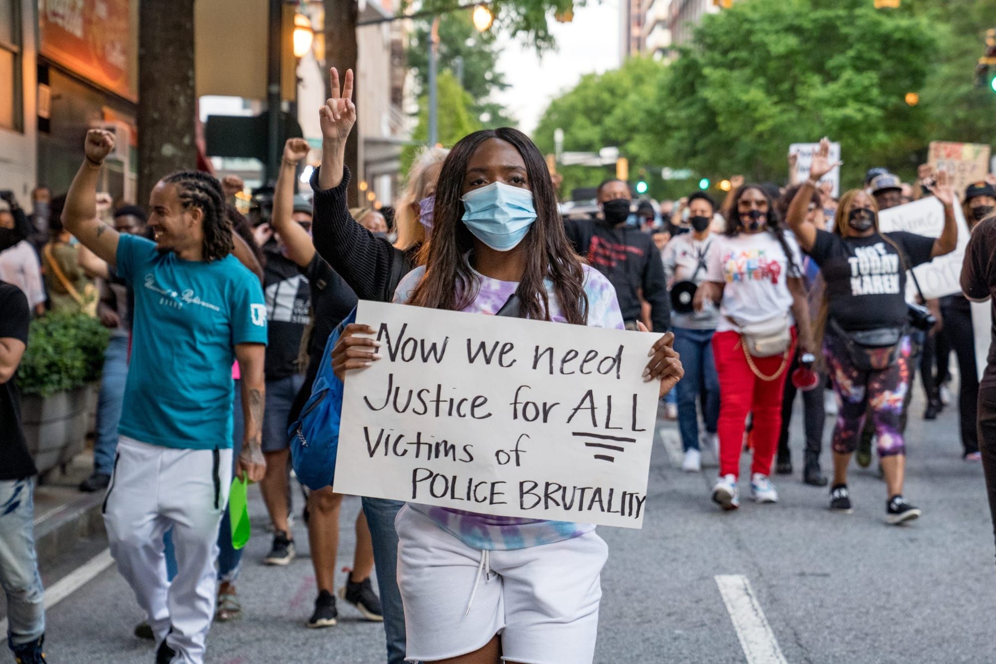 protesters walk in the street, and a Black femme wearing a mask holds a sign in the foreground saying "now we need justice fo