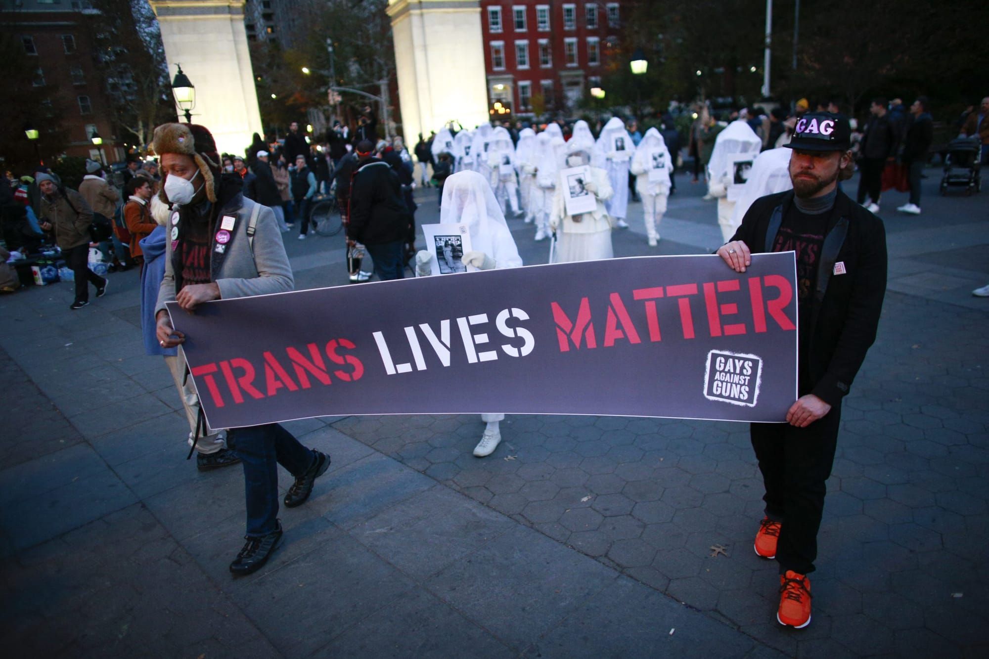 two people hold a banner reading "Trans Lives Matter" with a procession of about 15 people in white walking behind them