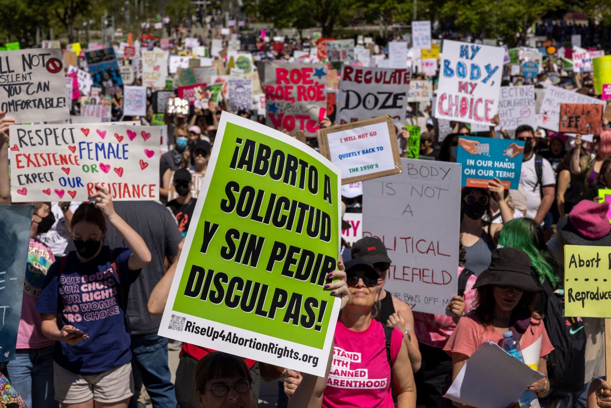 pro-abortion protestors hold signs outside. in the foreground, someone holds a sign reading "aborto a solicitud y sin pedir d