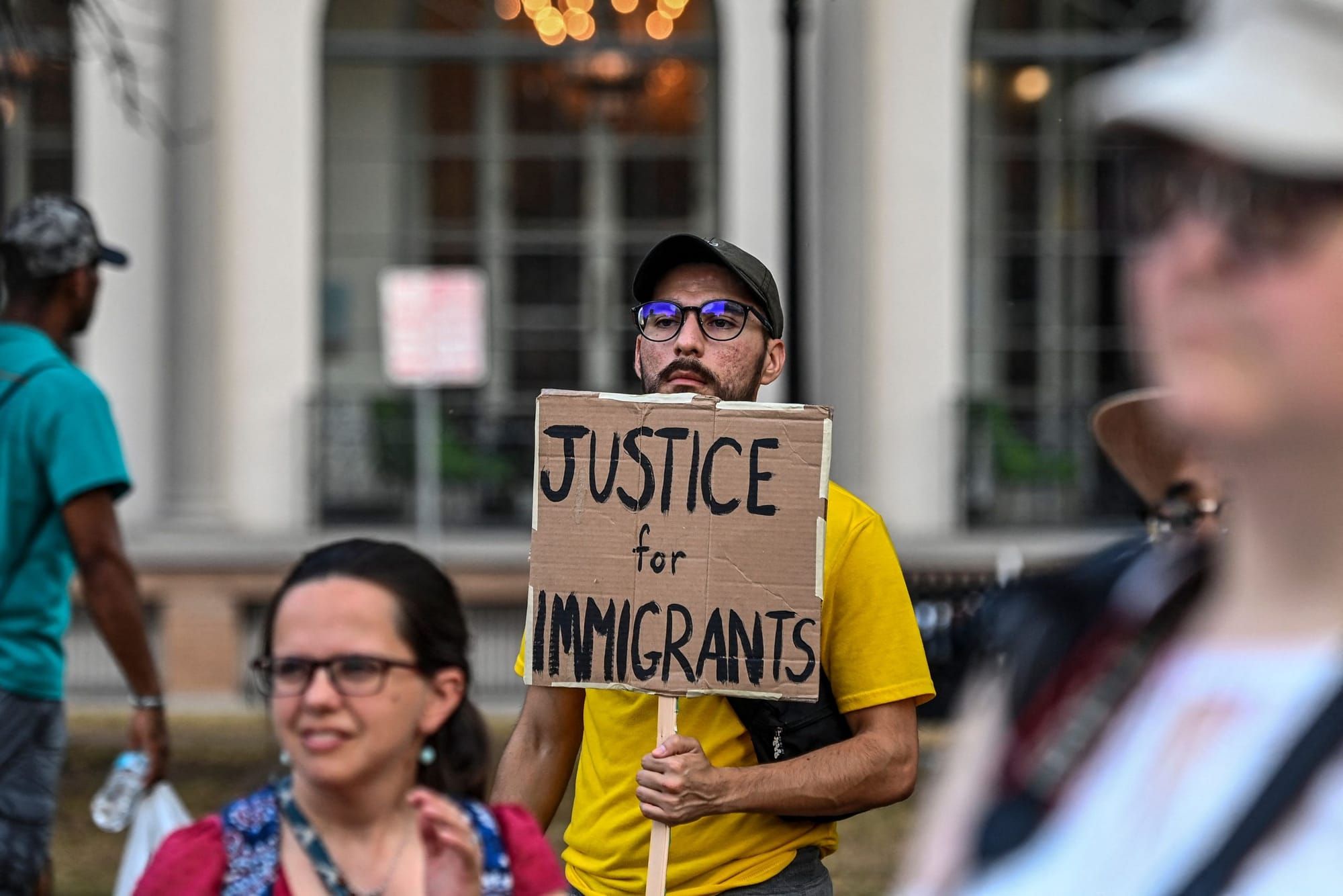 a person with a black mustache wearing a black baseball cap and yellow T-shirt holds a sign reading "Justice for Immigrants"