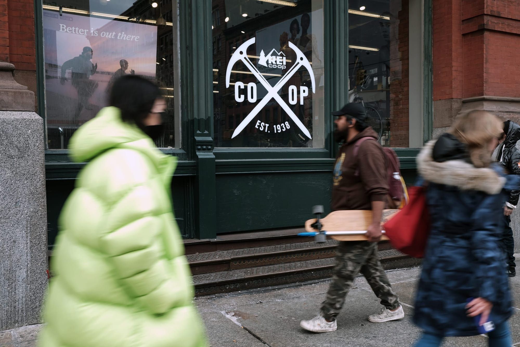 people in winter jackets walk on the sidewalk past the window of an REI Co-Op location