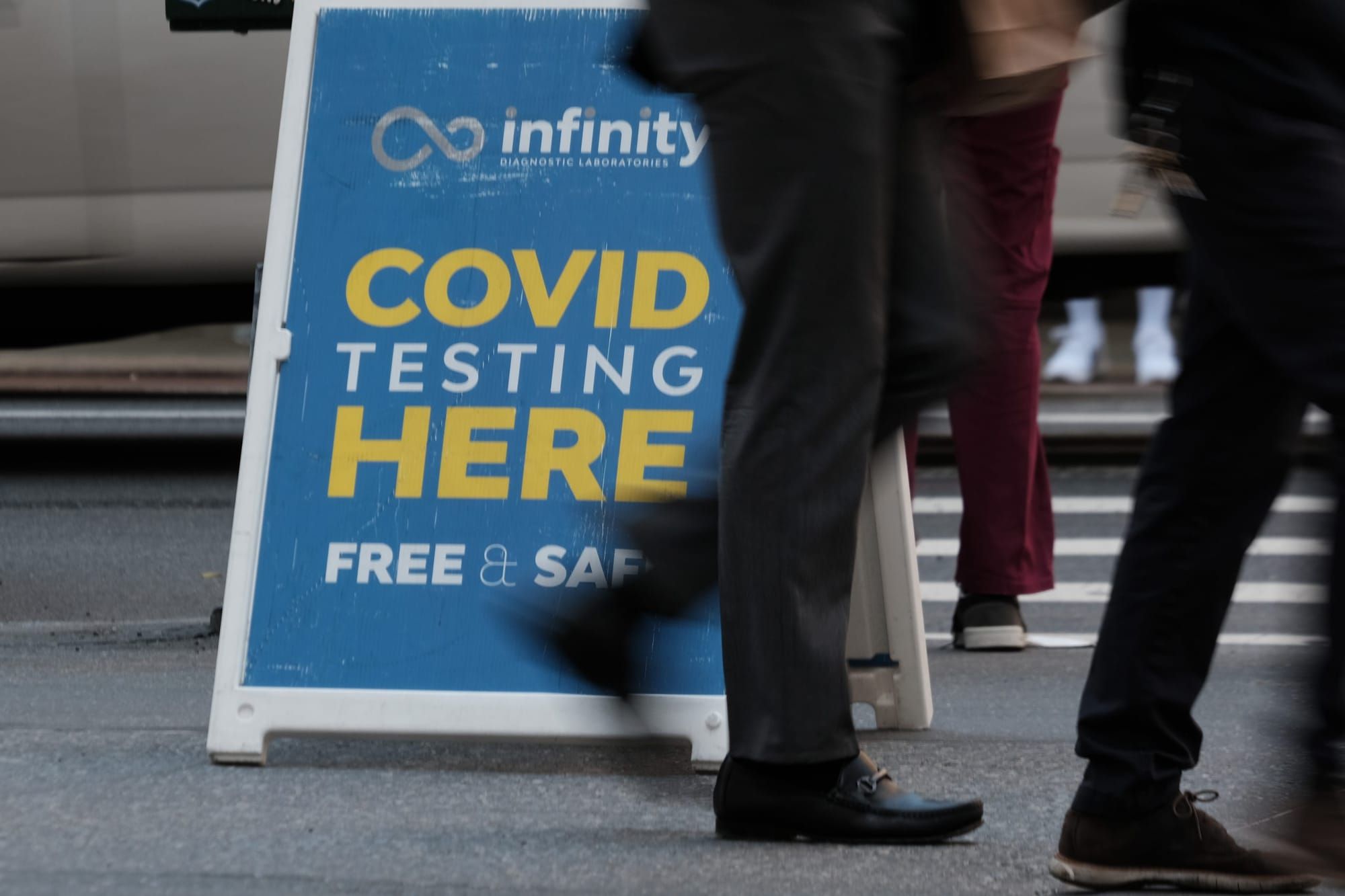 people walk past a blue sidewalk sign with yellow and white text reading "covid testing here free and safe"