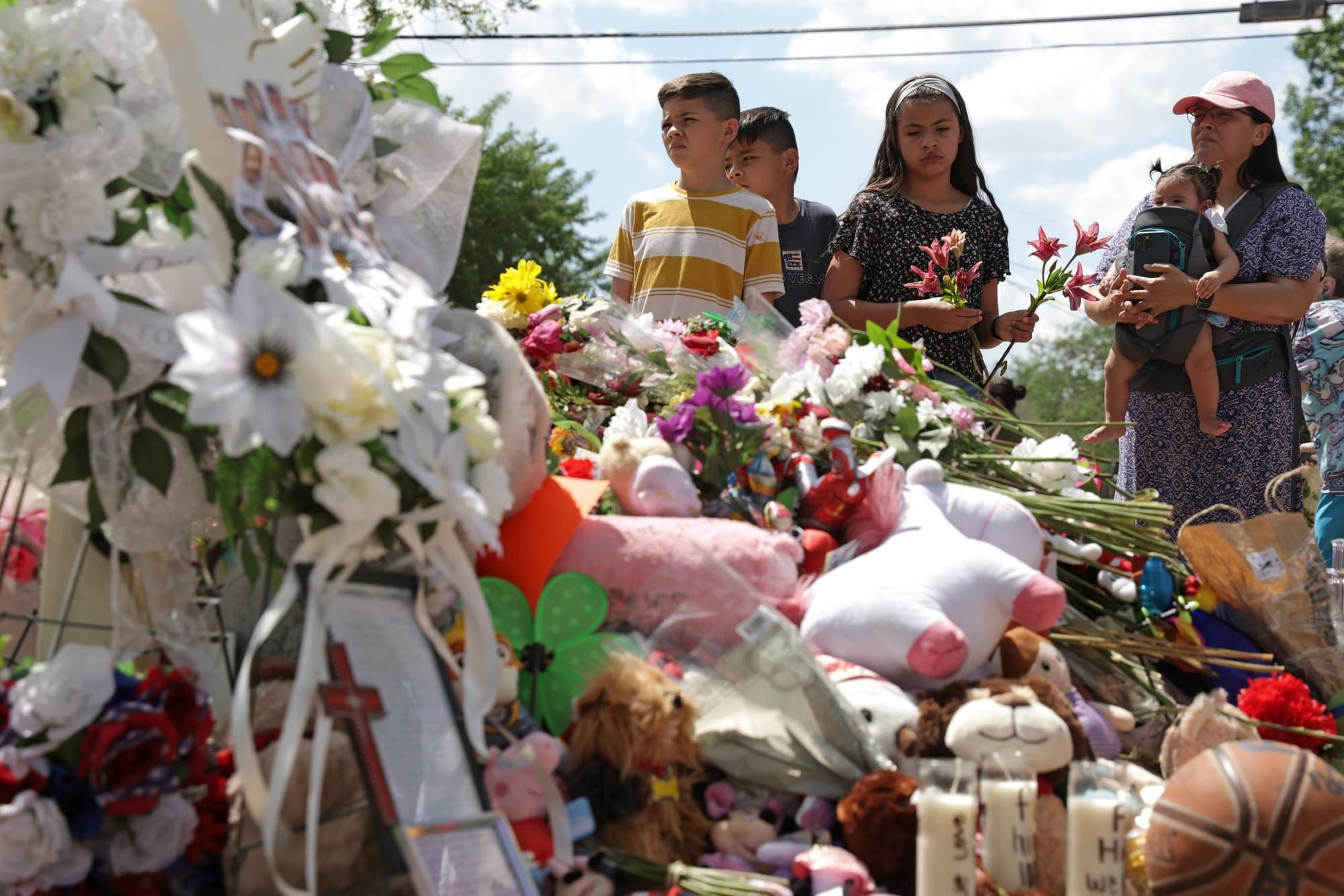 three young Latinx children stand with a Latinx woman holding a baby in front of a memorial for the victims of the Robb Eleme