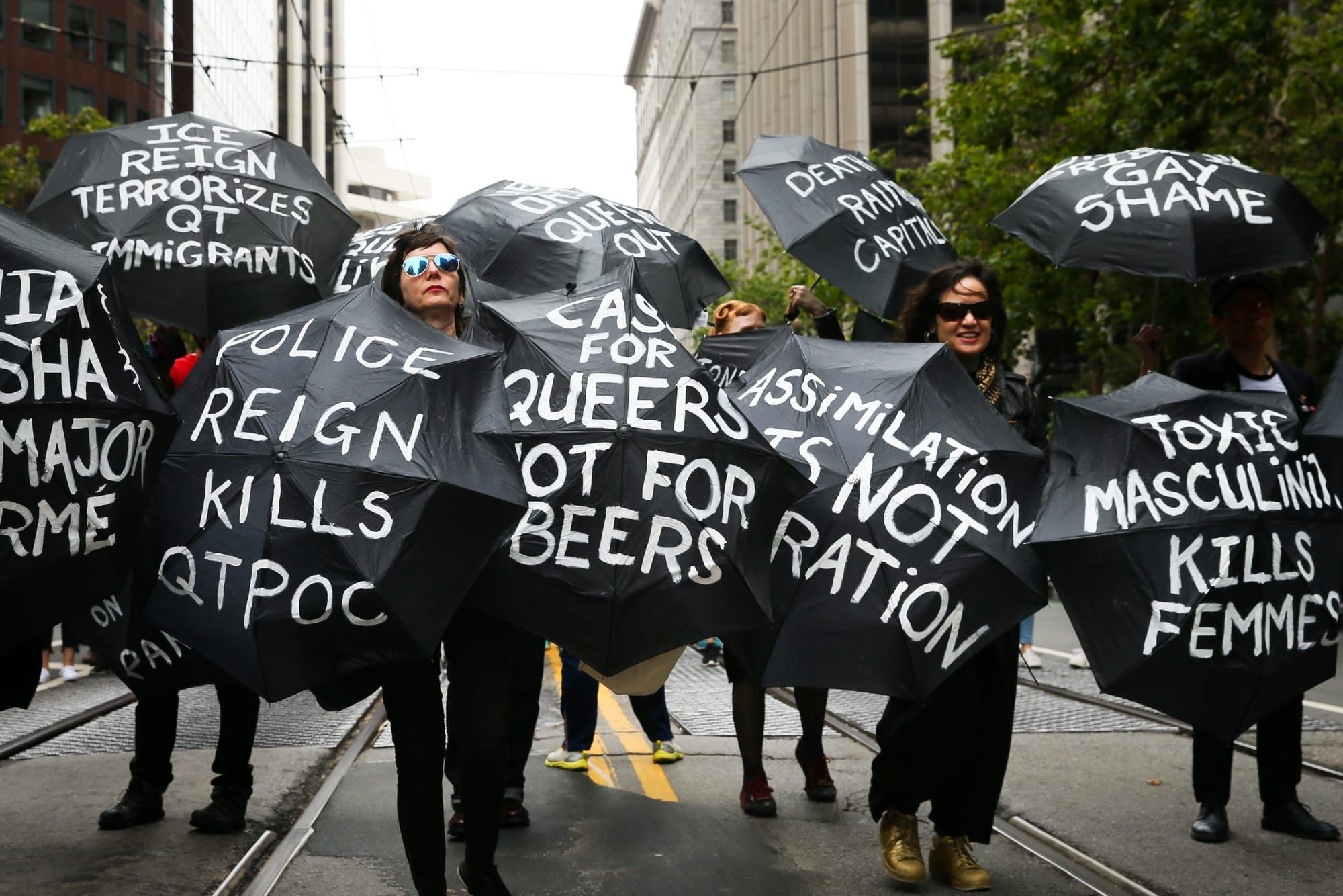 people attending Pride wear all black and carry black umbrellas with white text reading "Police reign kills QTPOC" and "ICE r