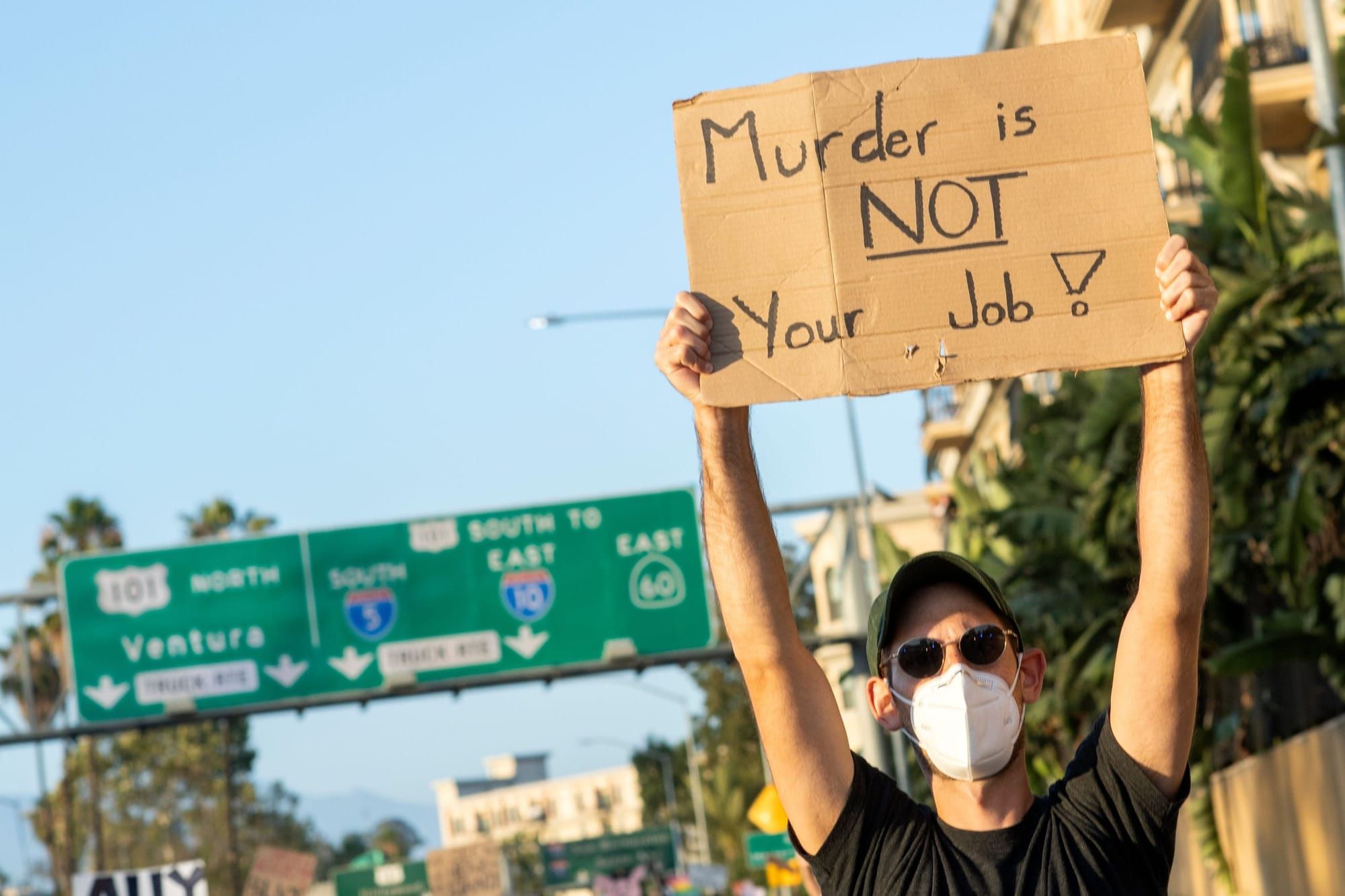 a person wearing a white medical mask holds up a cardboard sign reading "Murder is NOT your job!" while walking on a highway