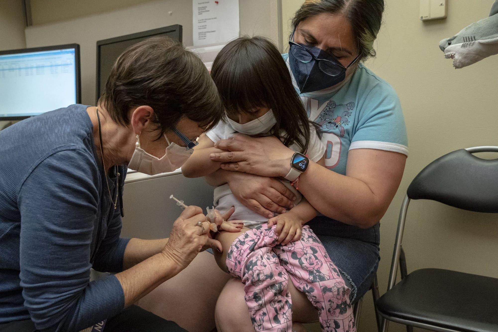 a female health care provider with short hair gives a COVID vaccine to a 2-year-old sitting in her mother's lap