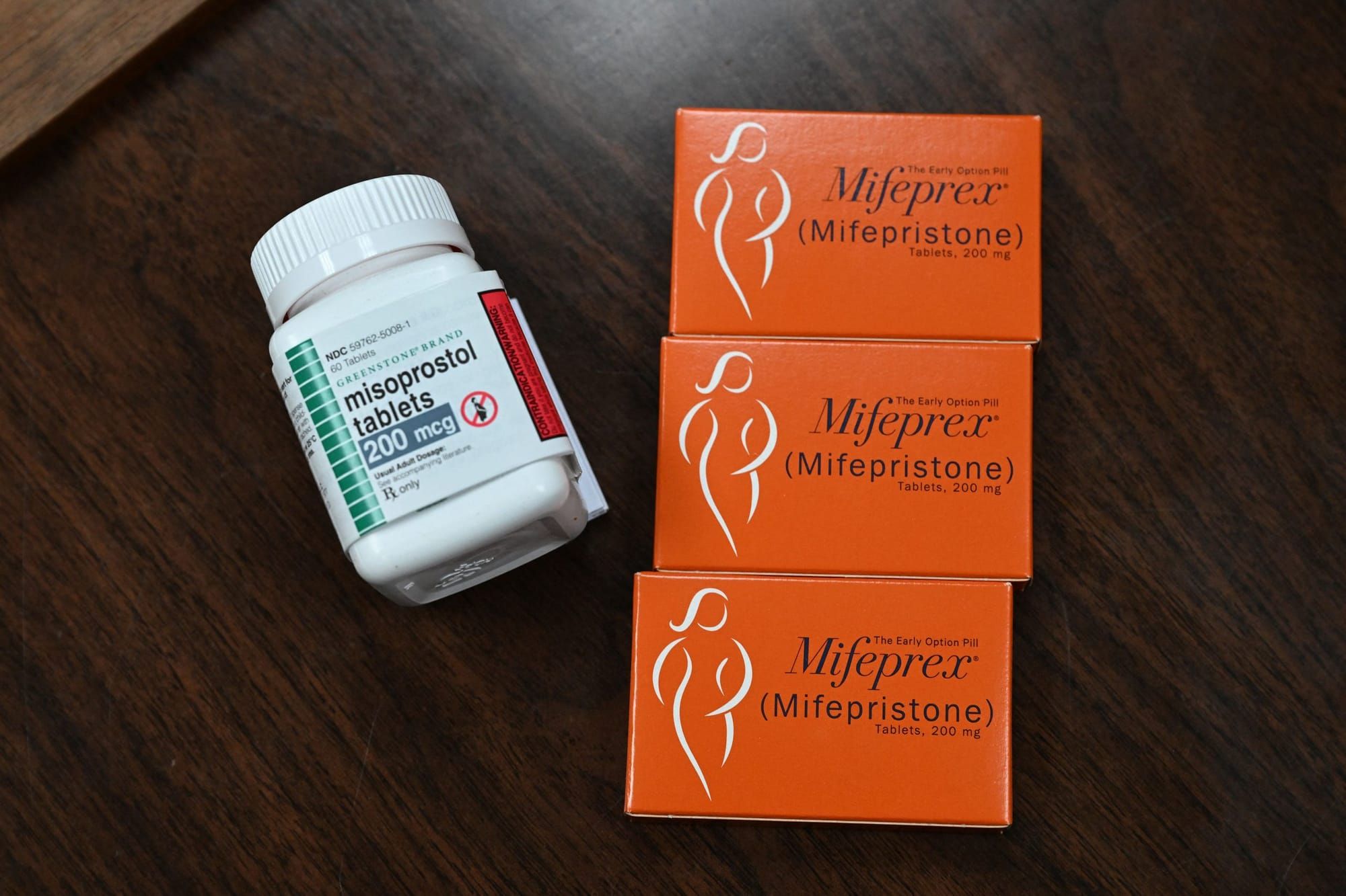 a white bottle of misoprostol on the left and three orange cardboard boxes of mifepristone sit on a dark brown wooden table