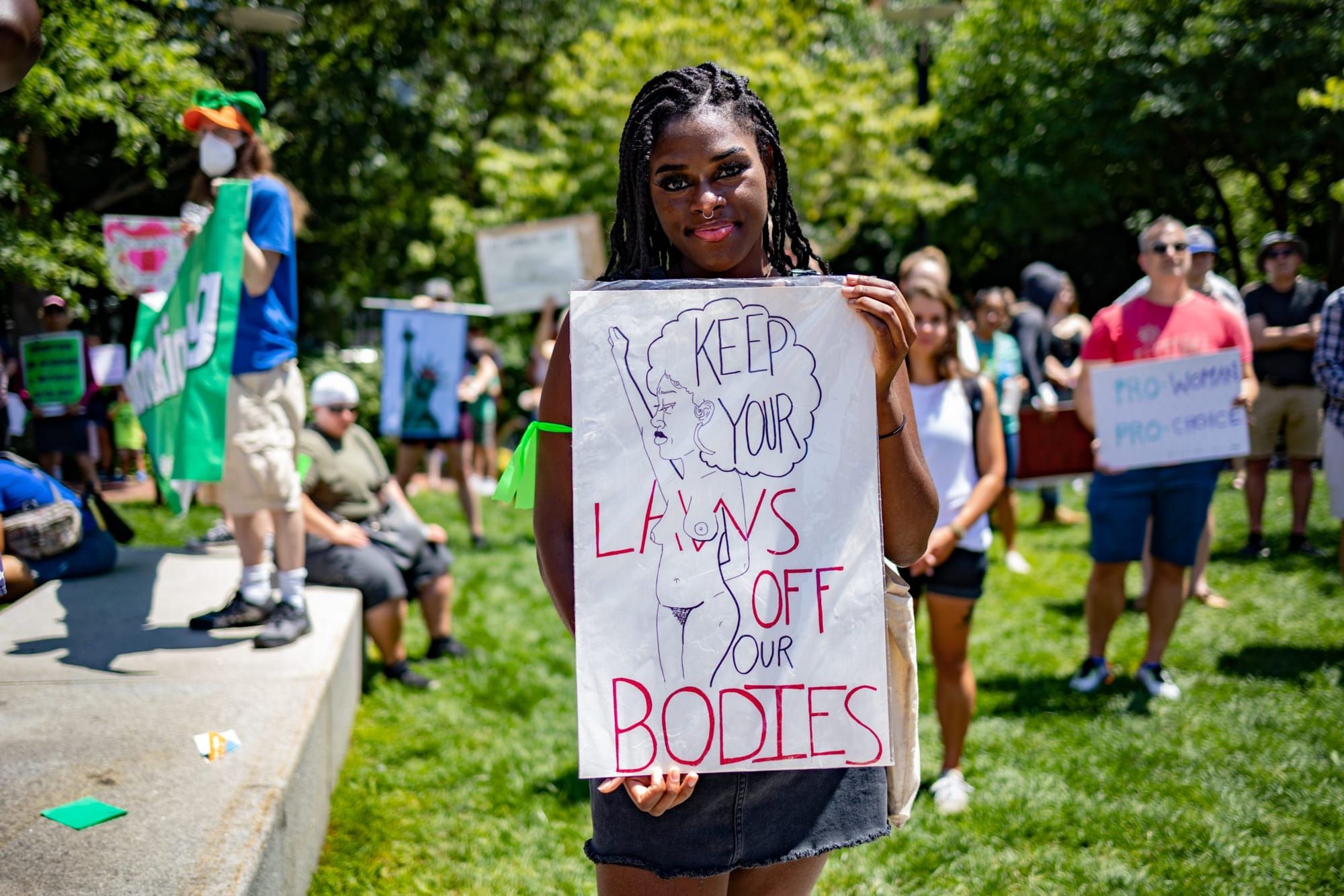 a Black femme holds a white poster saying "Keep your laws off our bodies" in black and pink ink