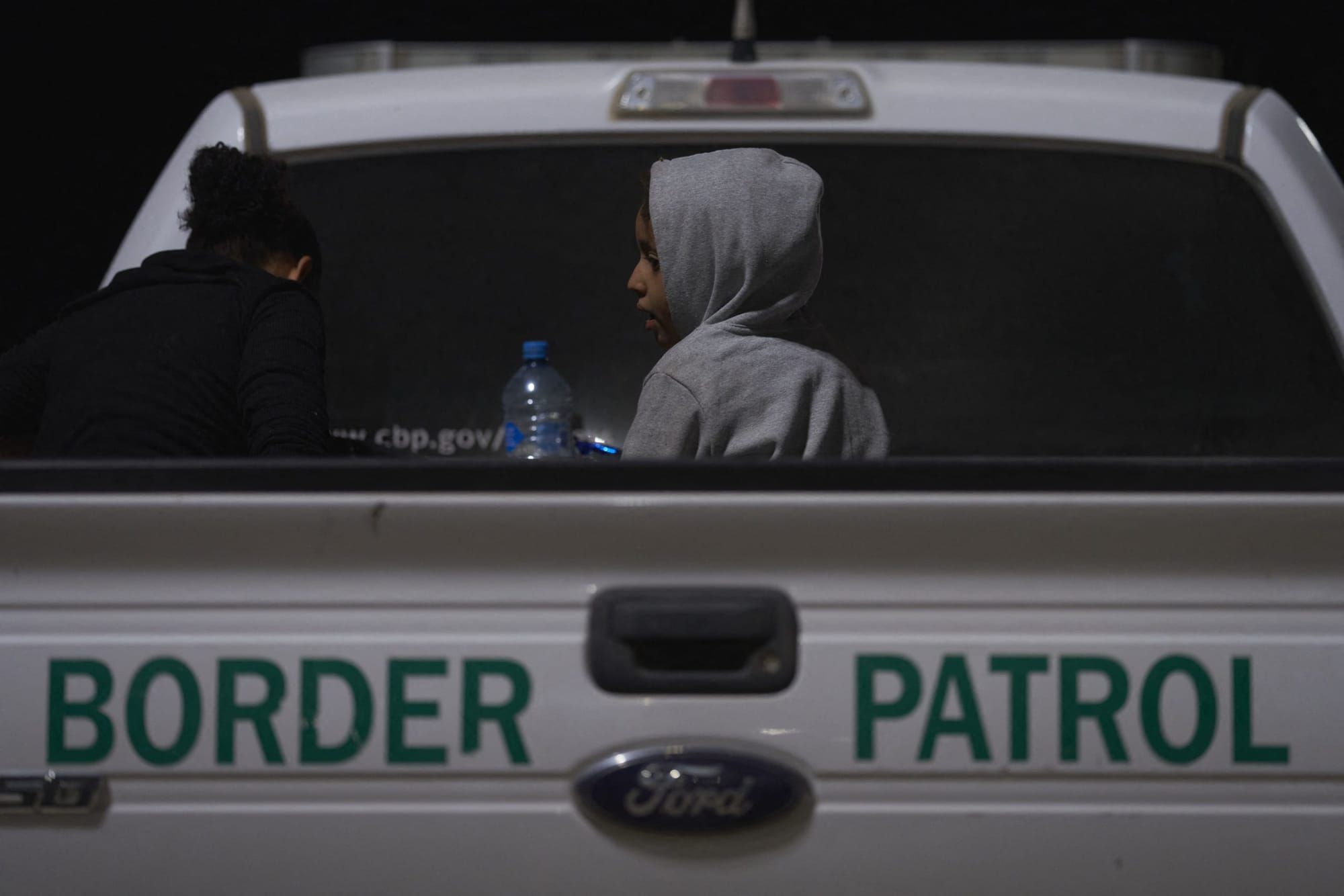 a child wearing a grey hoodie with the hood up sits in the bed of a white pick-up truck with the words "Border Patrol" writte