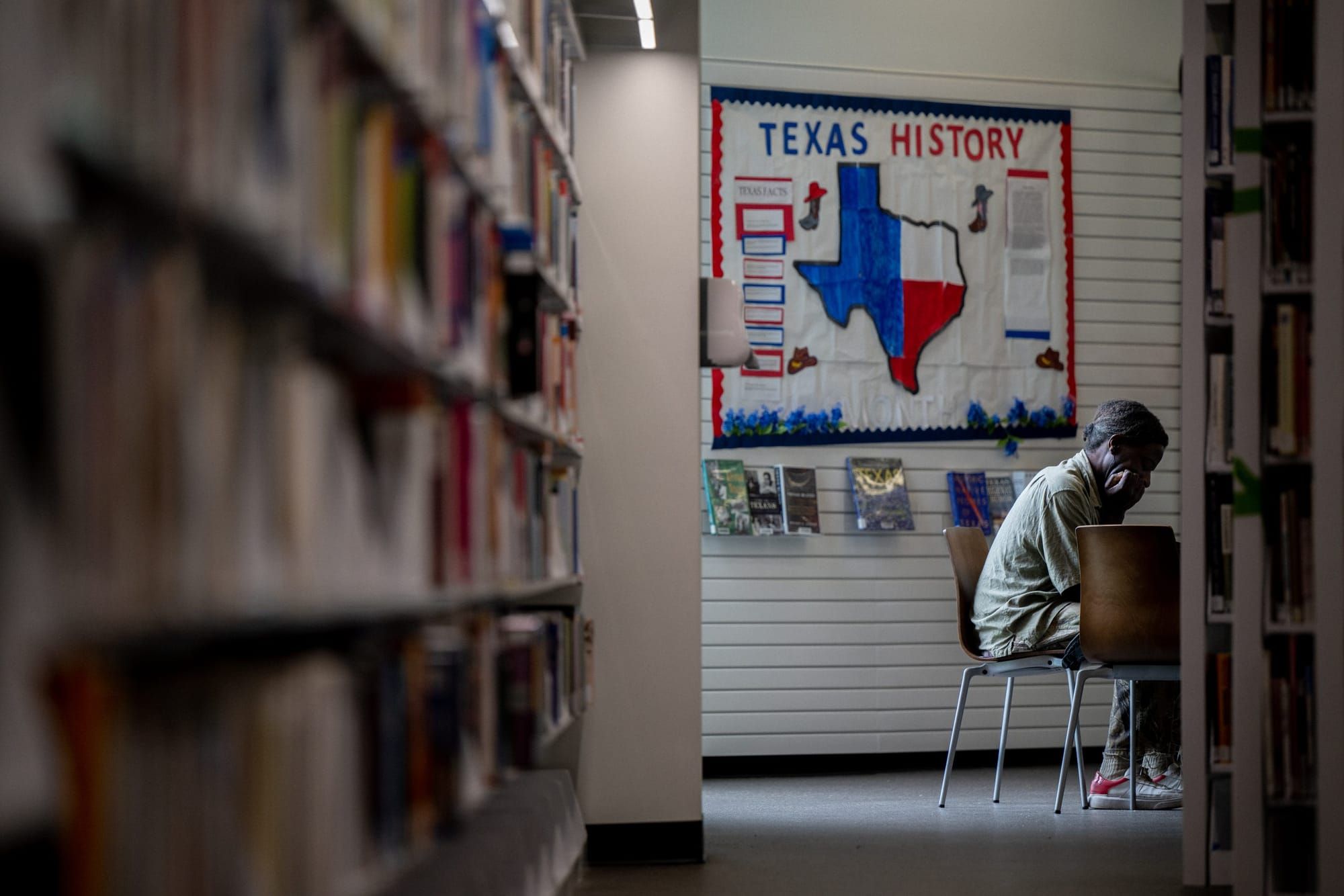 a student sits at a table in the background with a shelf of books blurred in the foreground. above and behind the student is