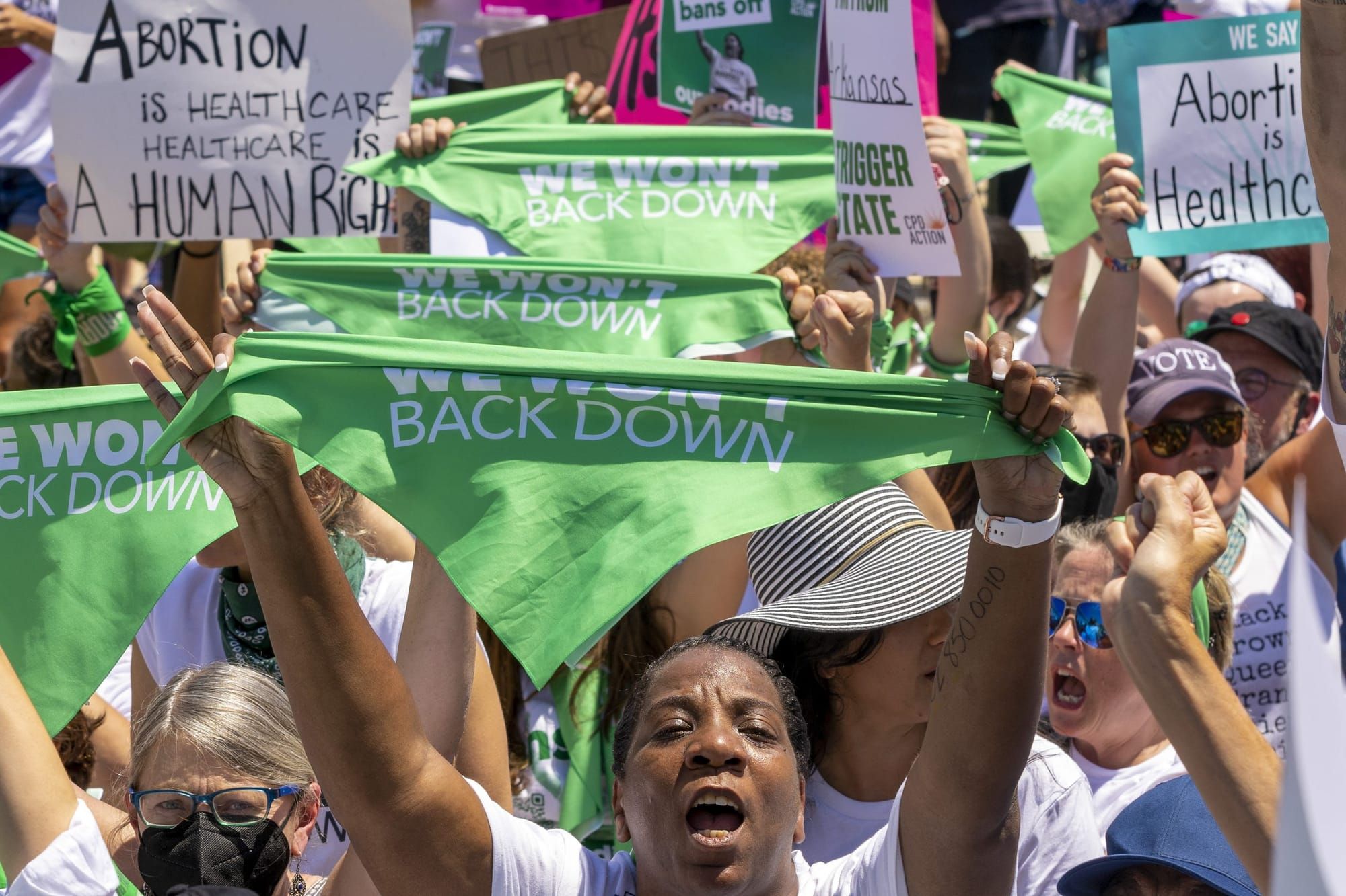 pro-abortion protesters hold up green bandanas that read "we won't back down" in white san serif font