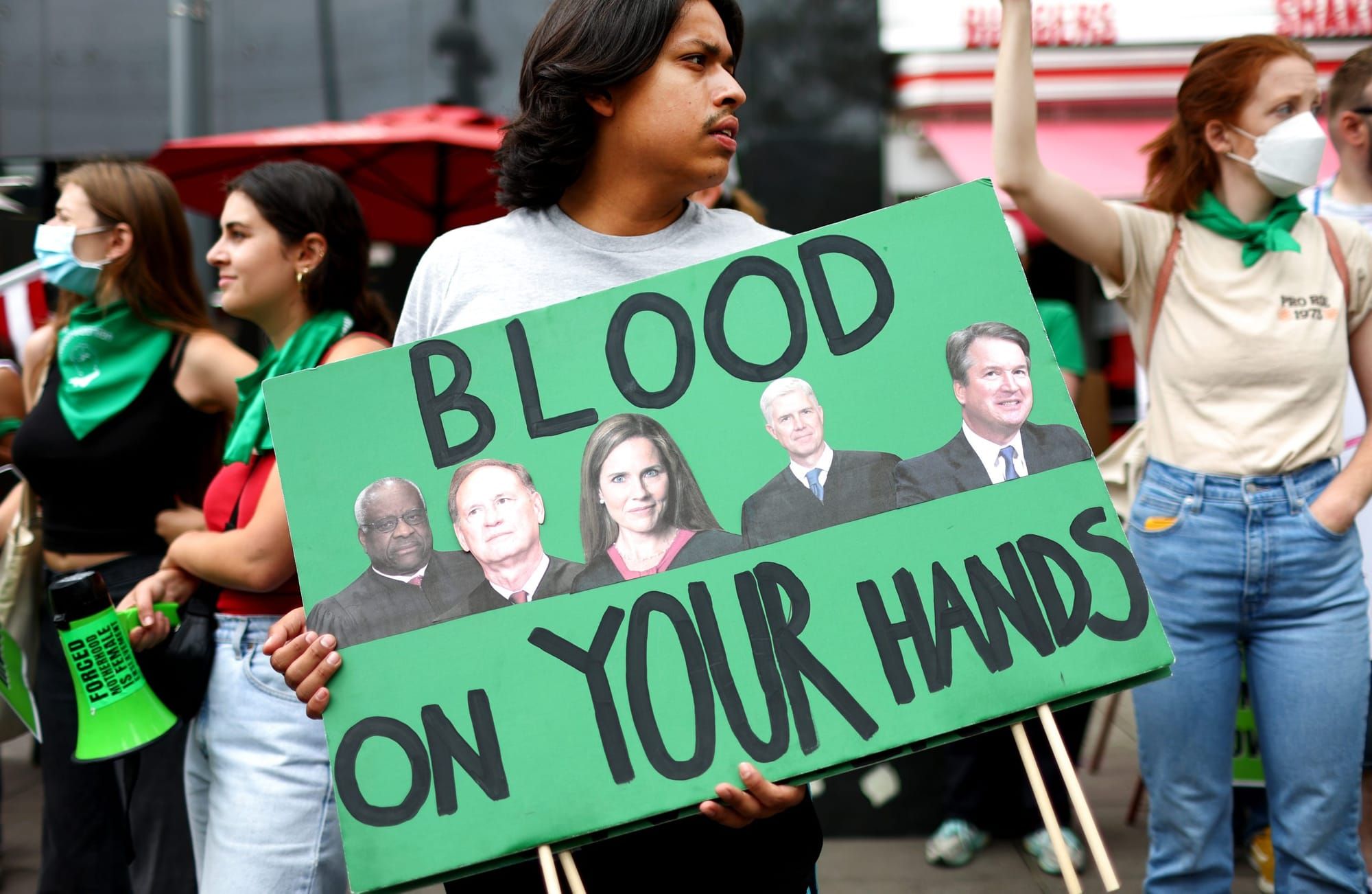 a pro-abortion protester holds a green sign with black-and-white cut-outs of the faces of Clarence Thomas, Samuel Alito, Amy