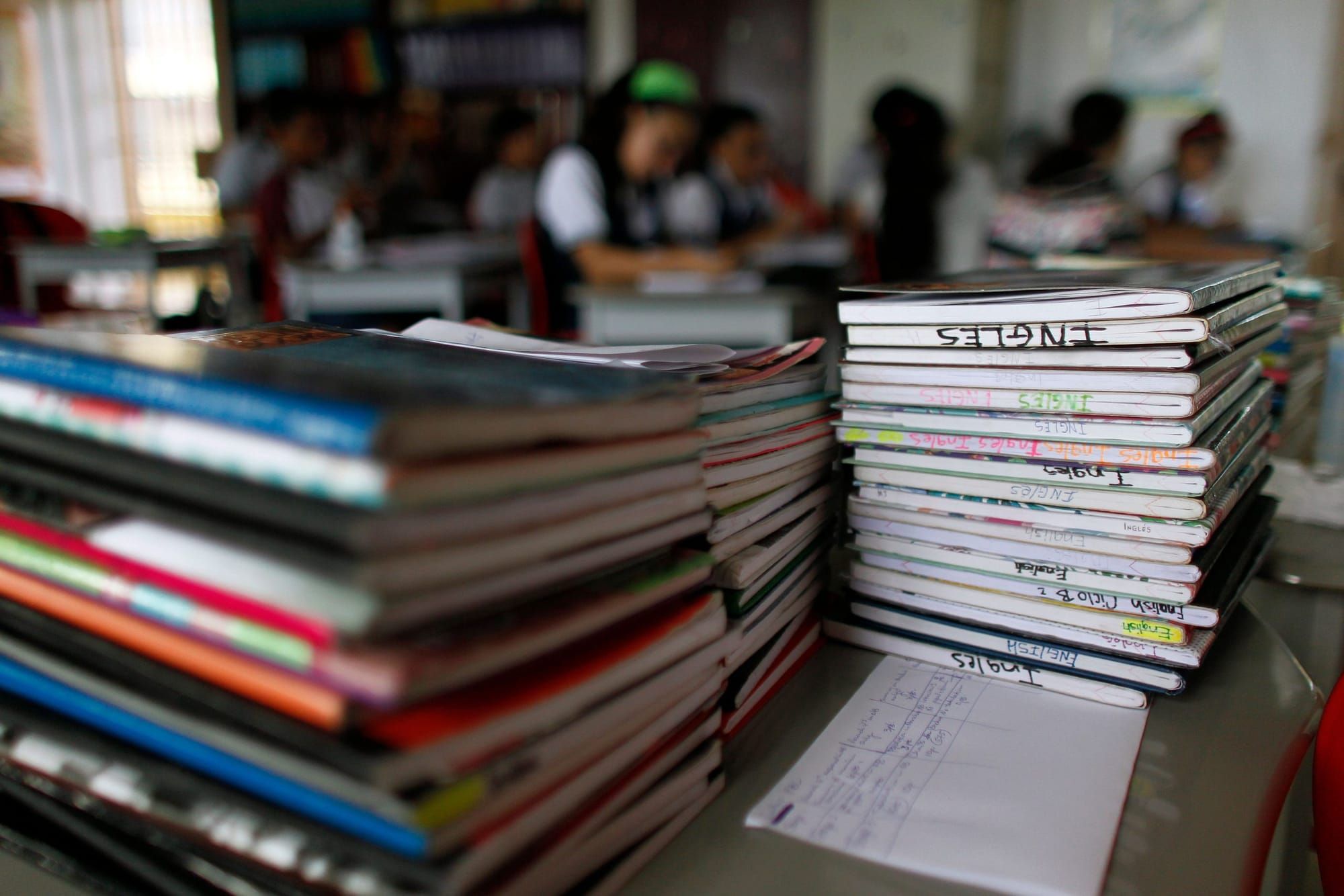 a stack of different colored notebooks in the foreground, with middle school students working at desks in a classroom in the