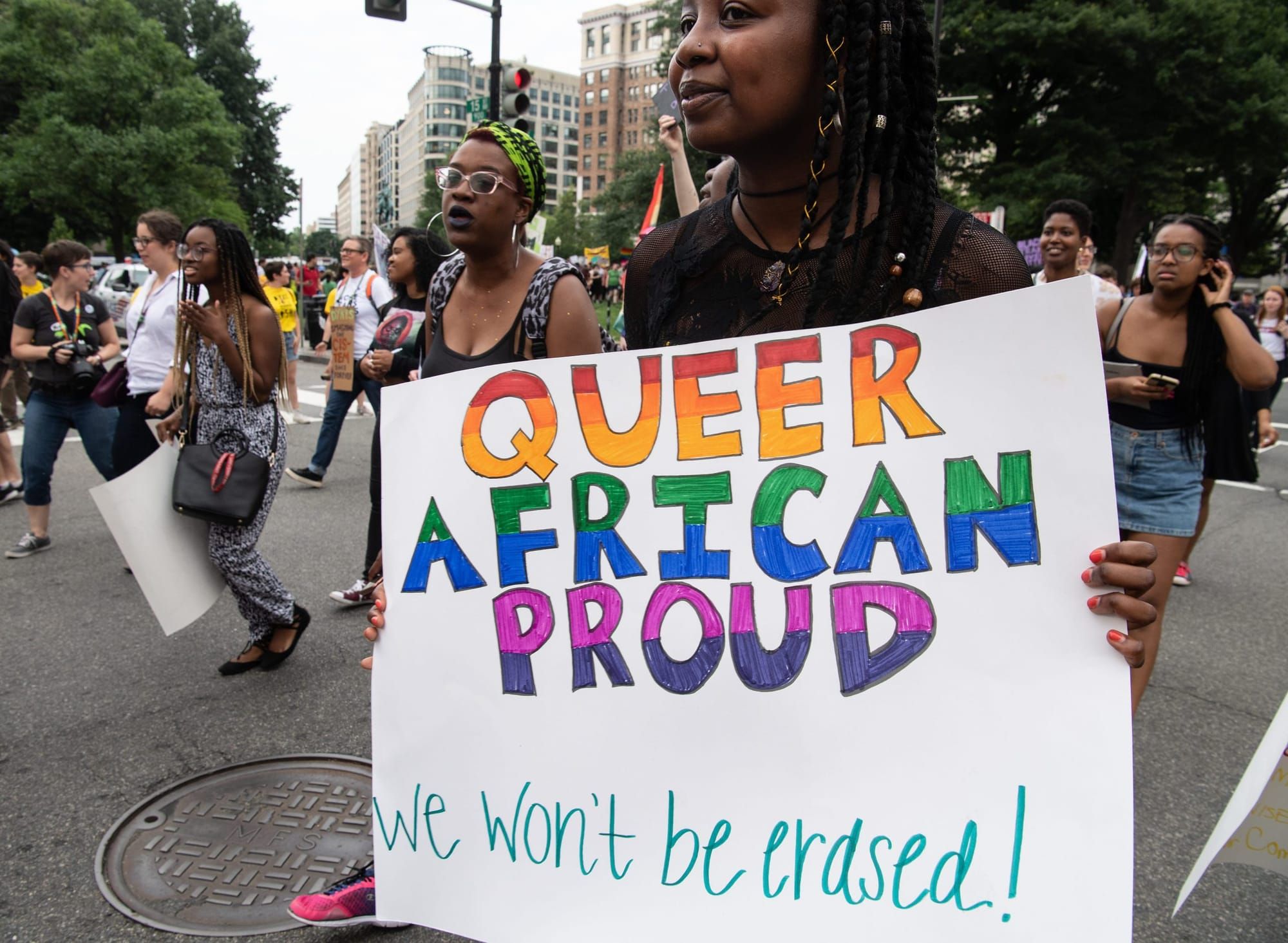 Black fem holds a white poster with rainbow lettering reading "Queer African Proud we won't be erased"