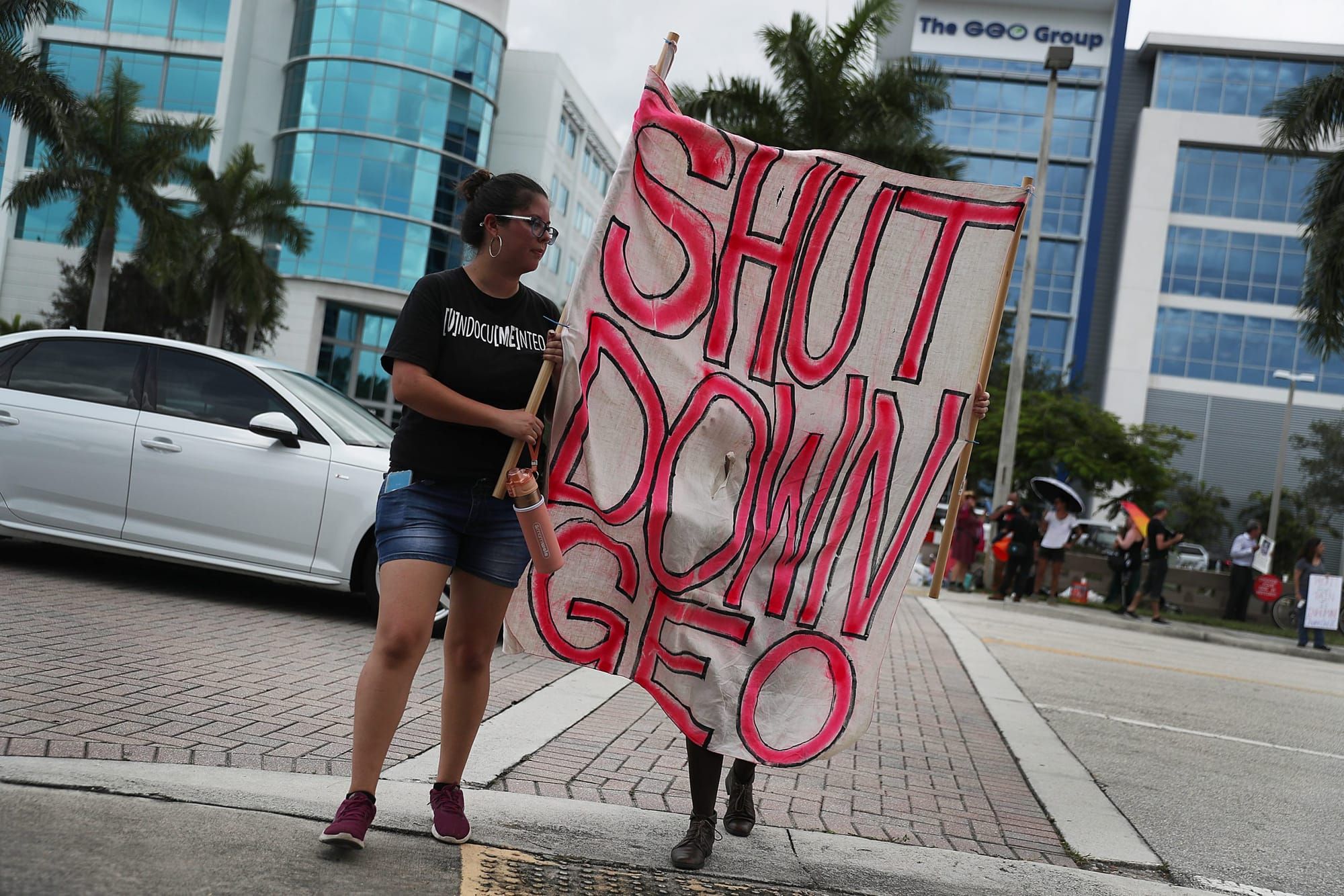 a femme person wearing short jeans and a black T-shirt reading "[U]ndocu[me]nted" helps hold a white poster with red letters