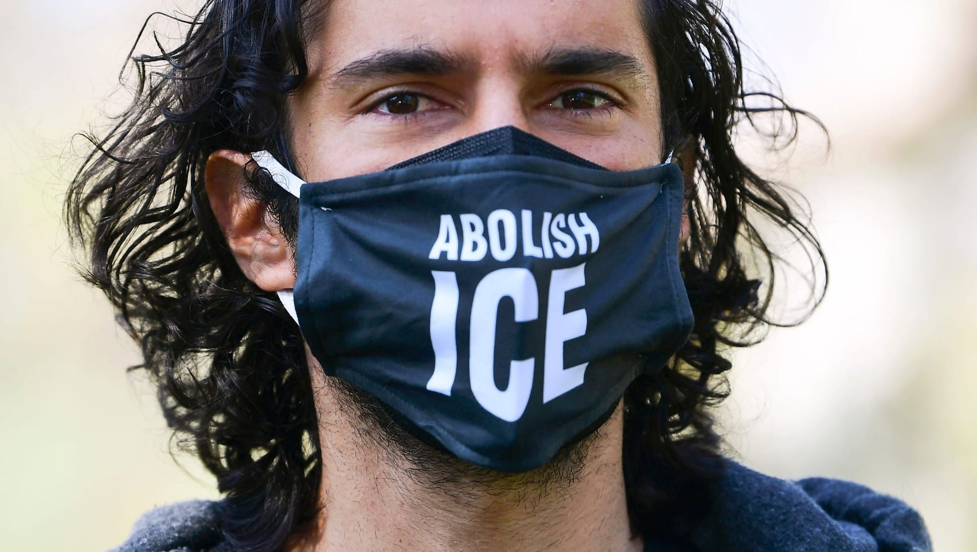 a person with short wavy hair down to their shoulders wears a black cloth mask with white letters reading "Abolish ICE"