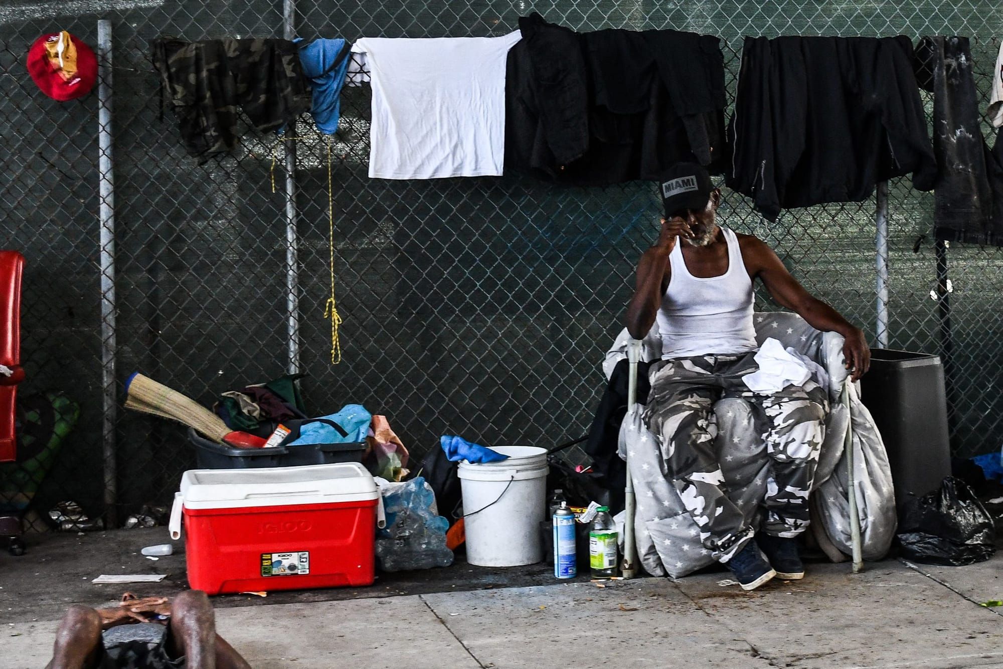 a black man in a white wifebeater and grey pants sits in a folding chair on a sidwalk in front of a chainlink fence wit ha co