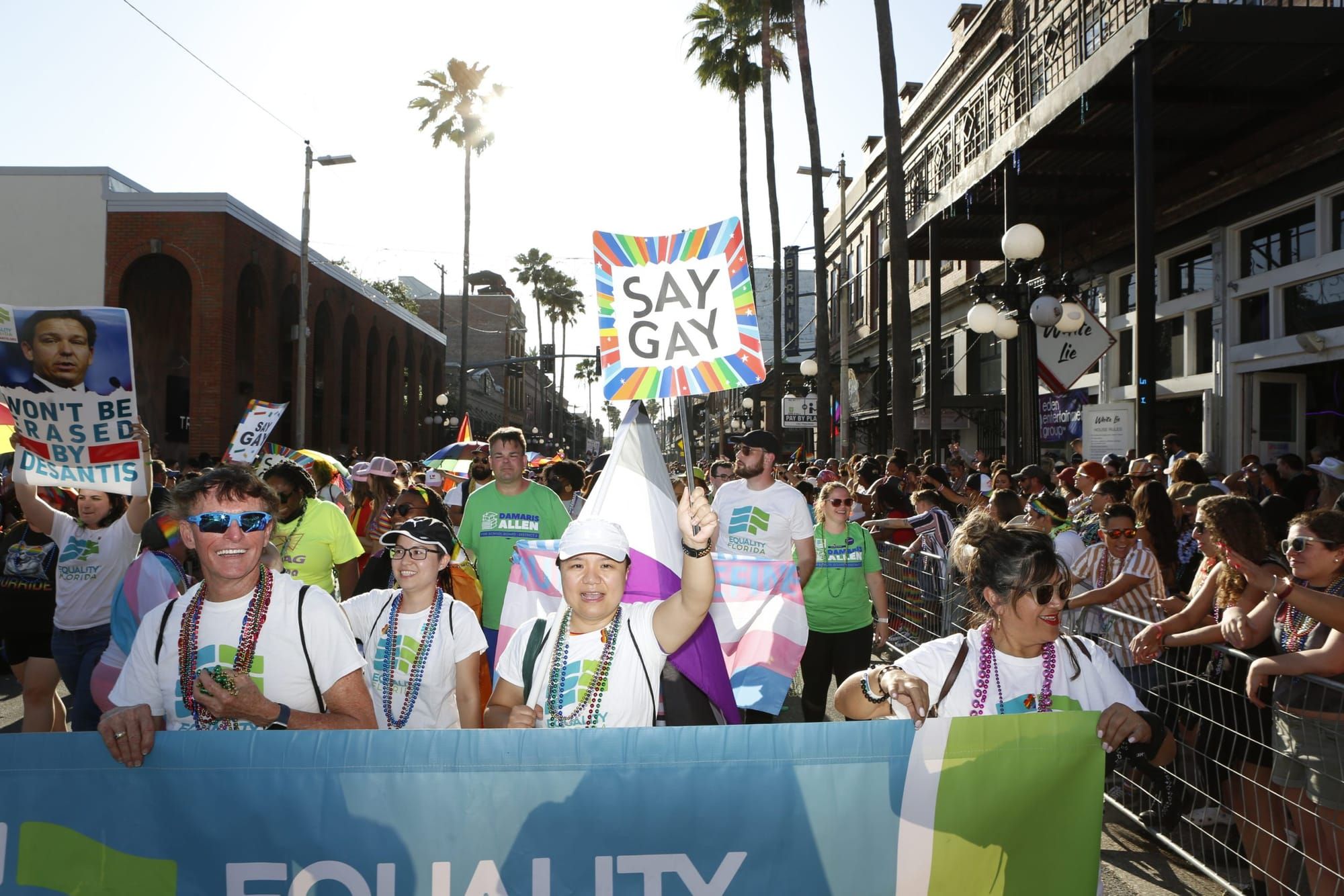 LGBTQ+ people and allies march in a parade while wearing white T-shirts with the text "Equality Florida" in blue and green. T