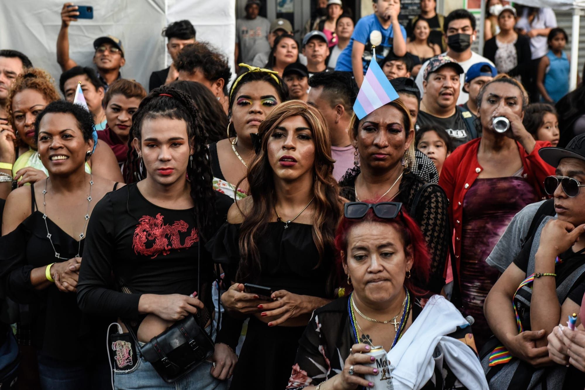 A crowd of Latinx people, some holding trans Pride flags, stands facing the camera