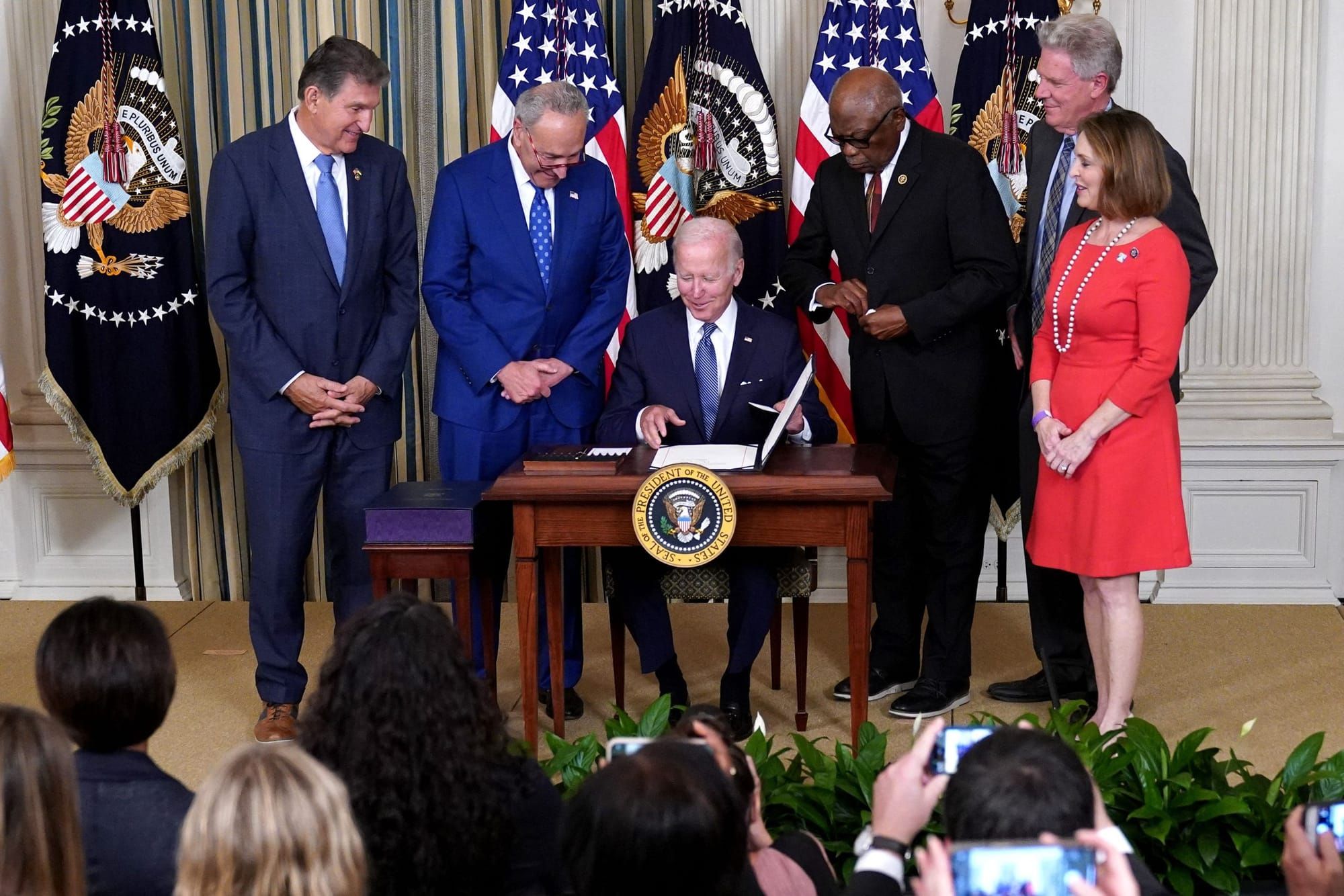 president joe biden wears a black suit with a white shirt and blue tie as he sits at a small wooden desk and signs the Inflat