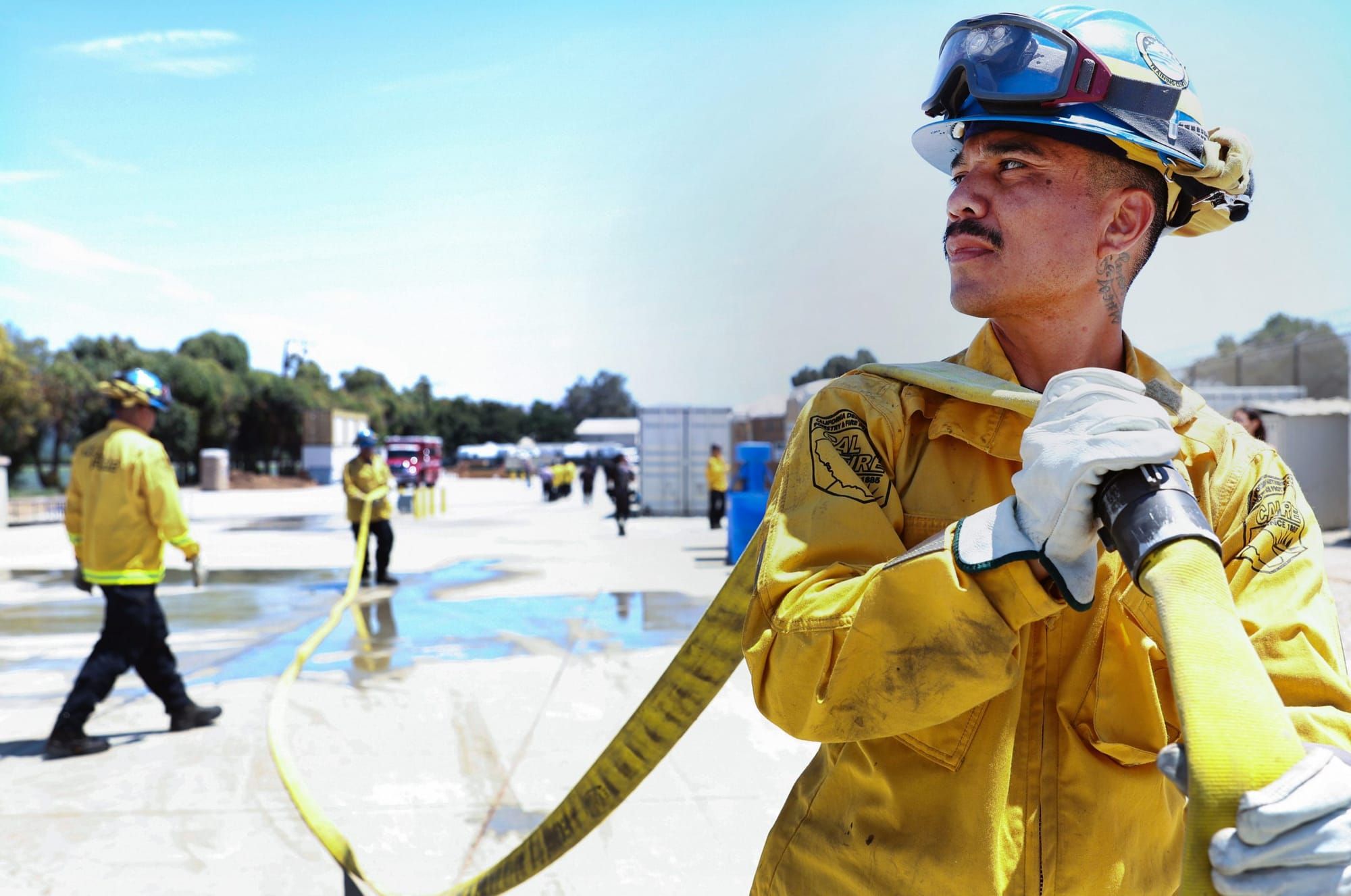 A Latino man with a mustache wearing a bright yellow firefighter's uniform holds a hose over his shoulder and looks to the le