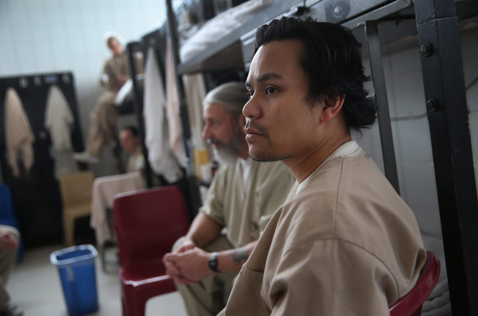 a Latino man in a tan inmates uniform sits in the foreground in front of other male inmates in the same outfit sitting on cha