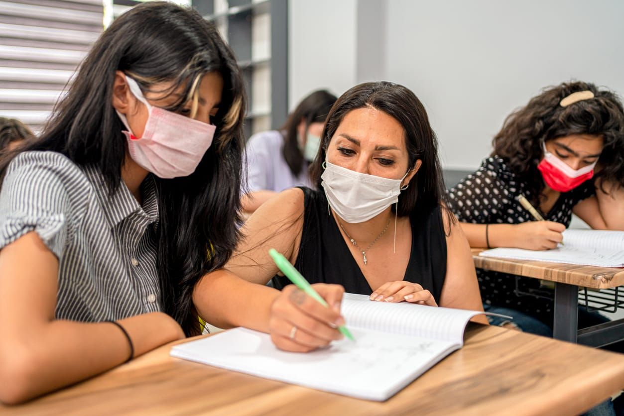 A Latinx teacher with long black hair wears a mask while leading a class of elementary school children. She is kneeling by th