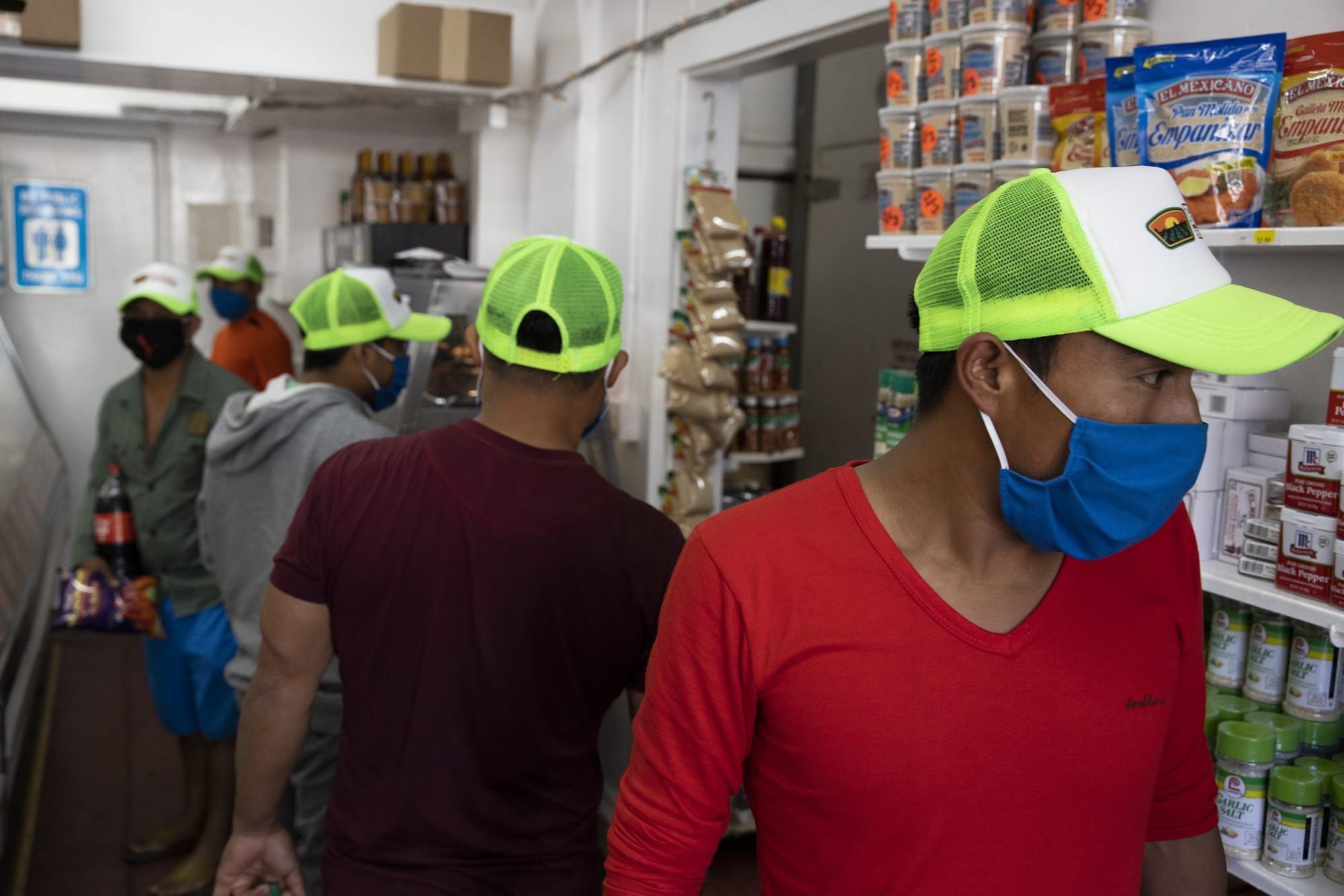 Latino workers in t-shirts and casual long-sleeve shirts wear cloth masks and neon green baseball caps as they walk through a
