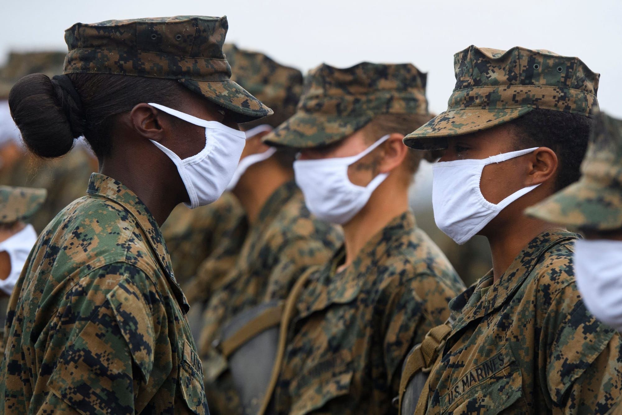 female marines stand in line in uniforms facing to the left. they wear white cloth face masks over their noses and mouths