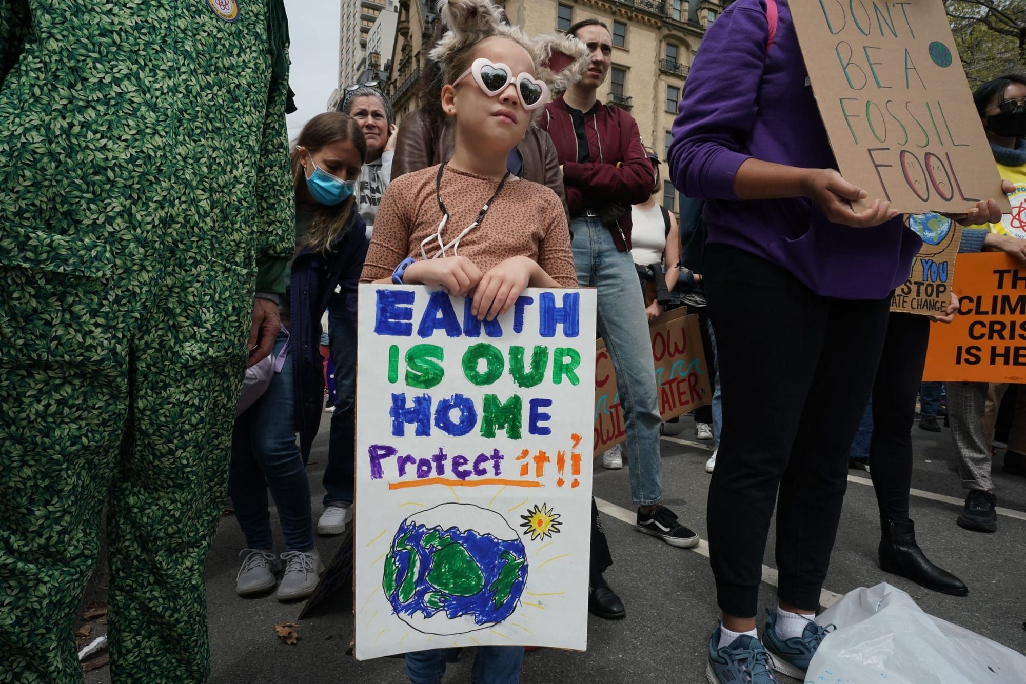 a young girl wearing white plastic heart-shaped sunglasses holds a white poster with blue and green text reading "Earth is ou