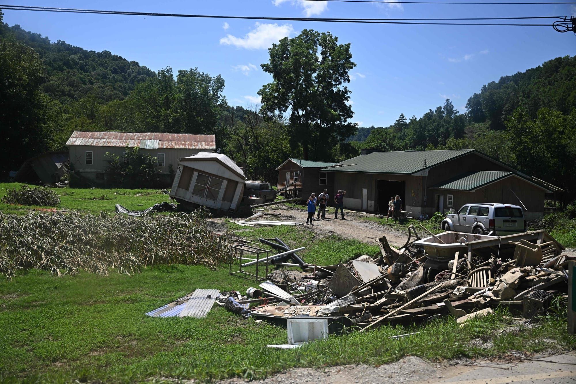a pile of wooden debris sits in the foreground on a grassy area. In the midground there is a tilted trailer and a pile of tre