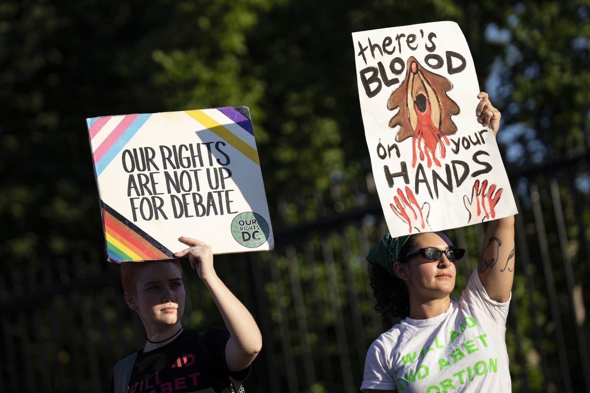 two people hold up signs in support of abortion. the one of the left reads "our rights are not up for debate" with Pride, tra