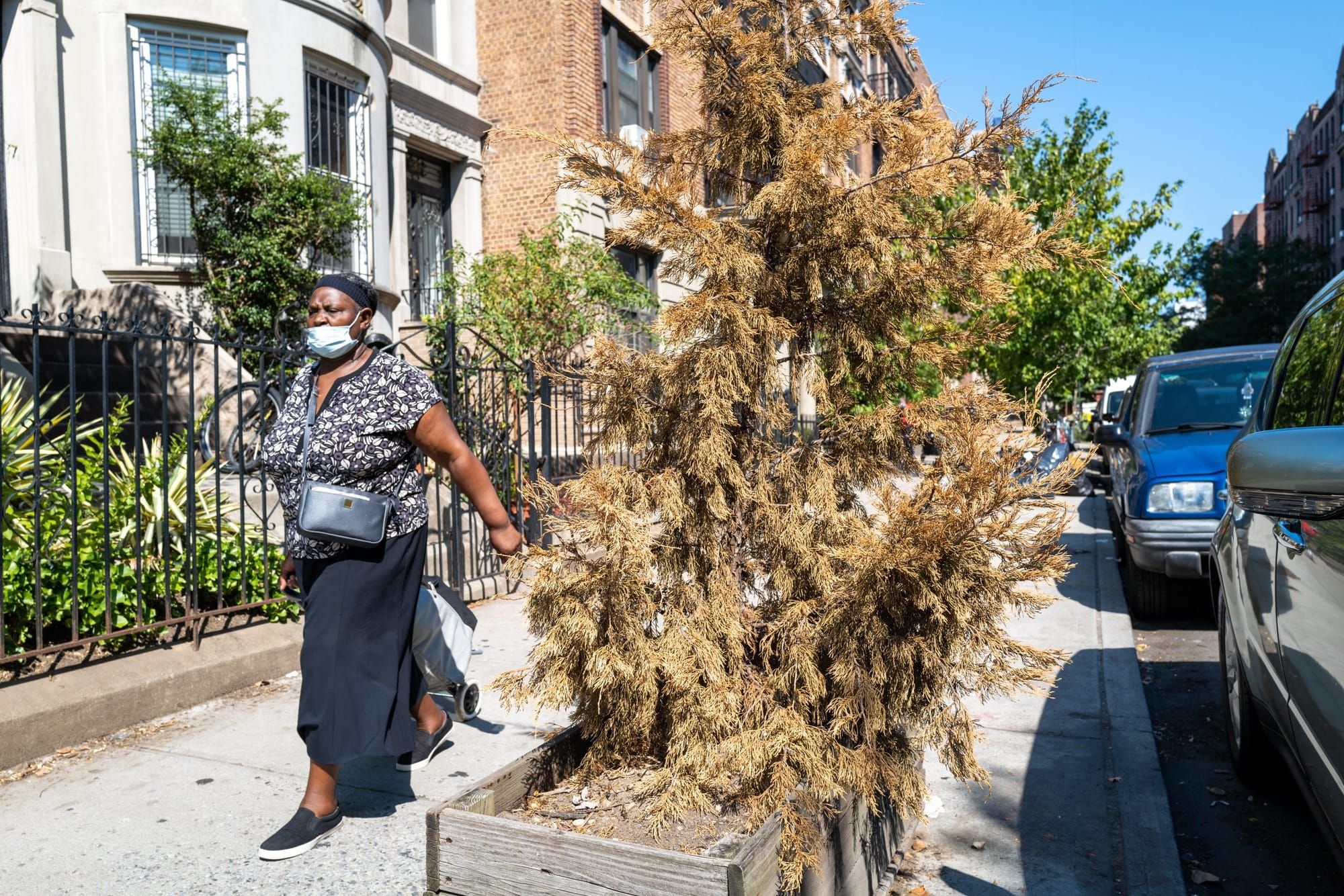 a Black woman wearing a blue disposable mask, a black-and-white patterned short-sleeved blouse, and a long black skirt walks