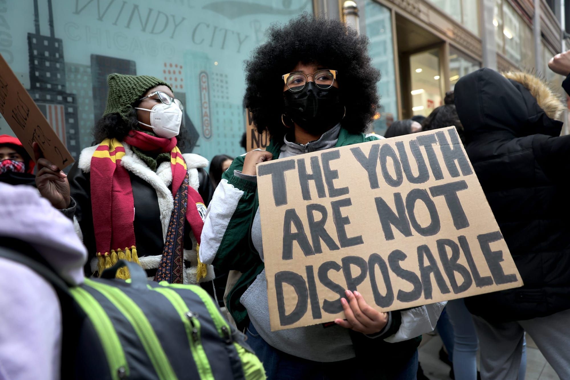 a Black woman with natural hair wearing a grey shirt and black jacket stands in a crowd. she holds a brown cardboard sign wit