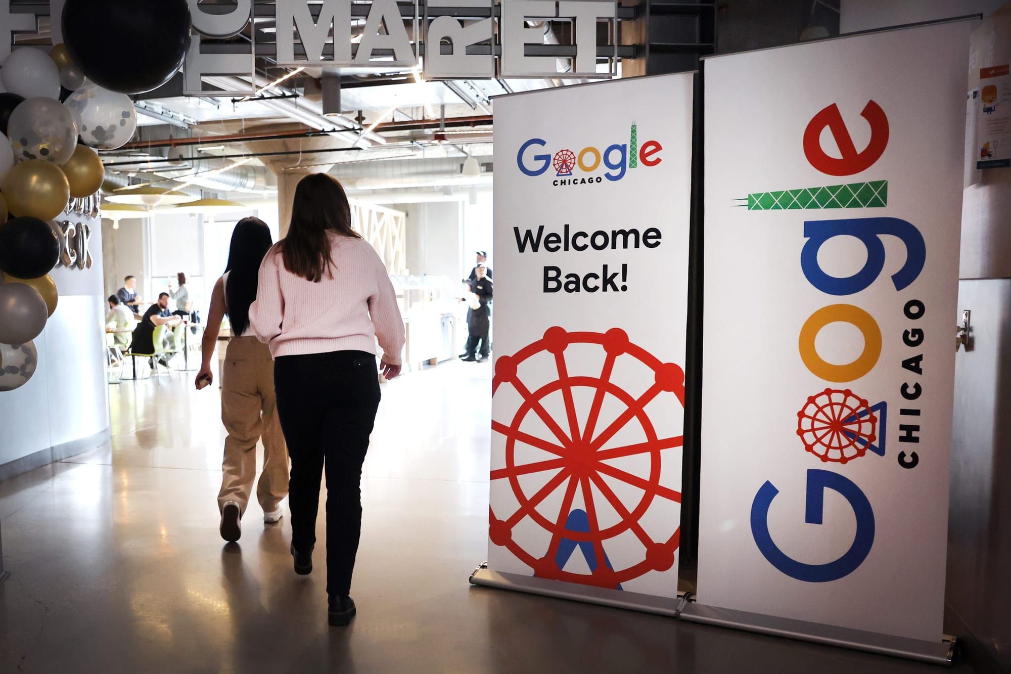 two women in business casual attire walk away from the camera past two large signs with Google logos and the text "welcome ba