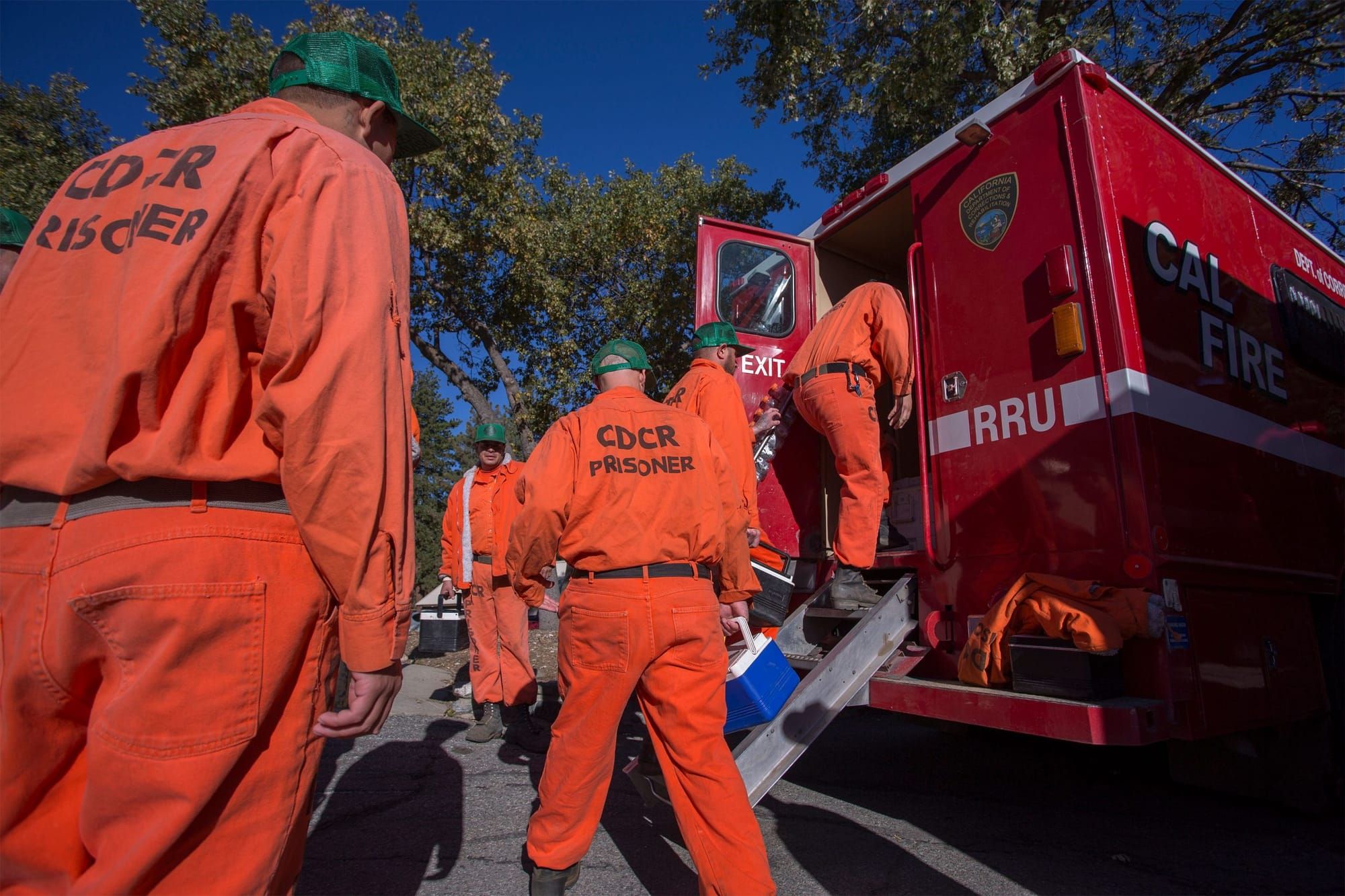 incarcerated firefighters wearing bright orange jumpsuits walk towards the back of a red firetruck. One firefighter is climbi