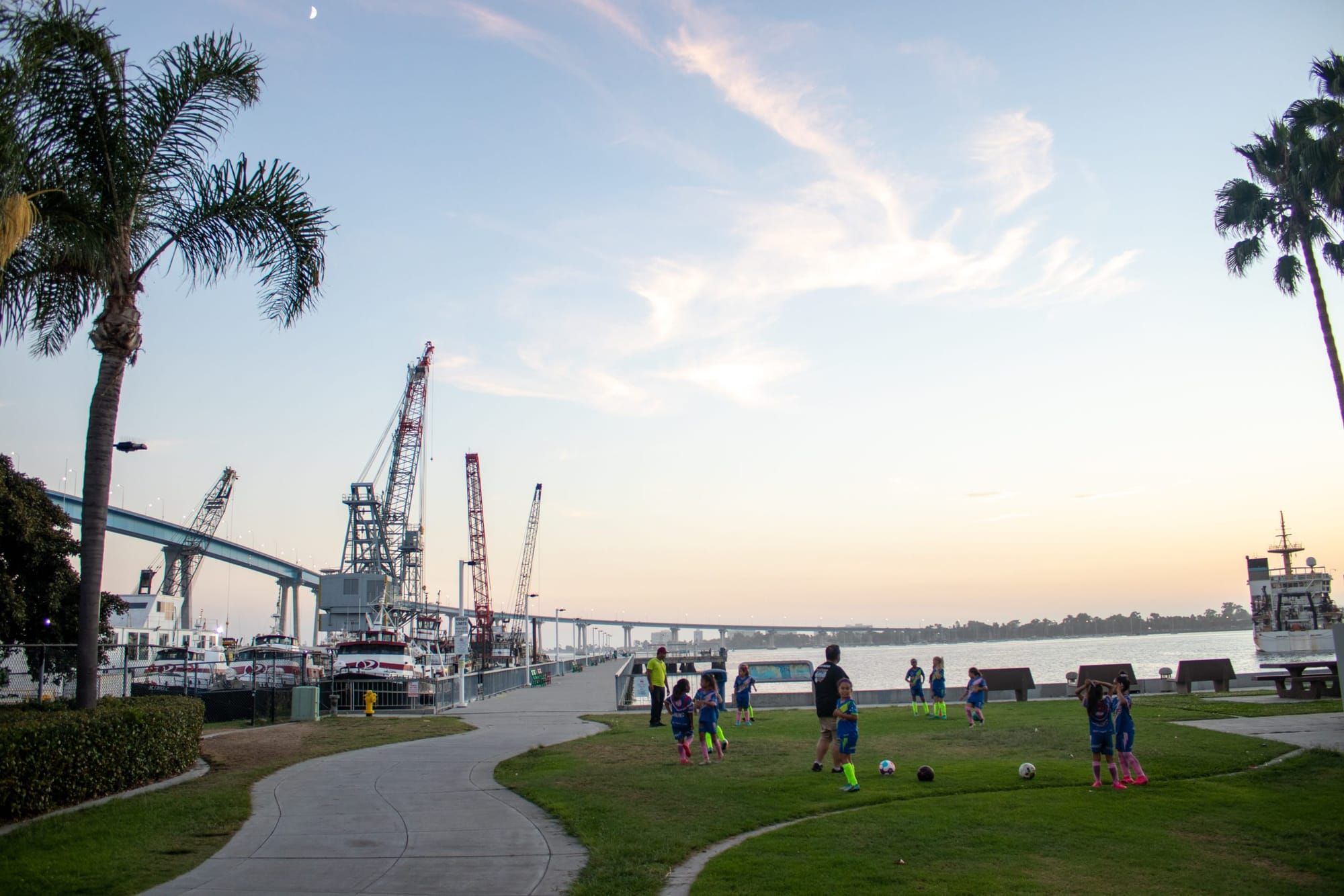 Latinx children play soccer with adults supervising on the field. In the background is a bay and cranes