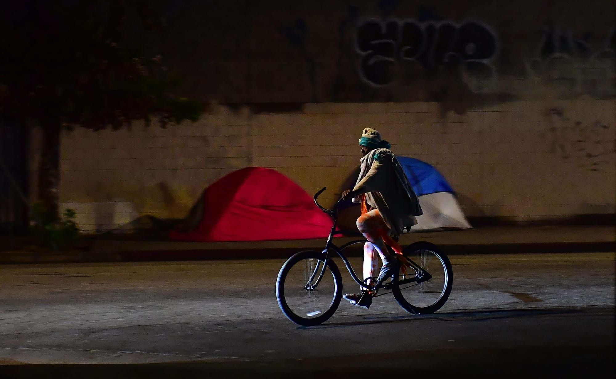 a Black person in a winter coat rides a bicycle past a homeless encampment