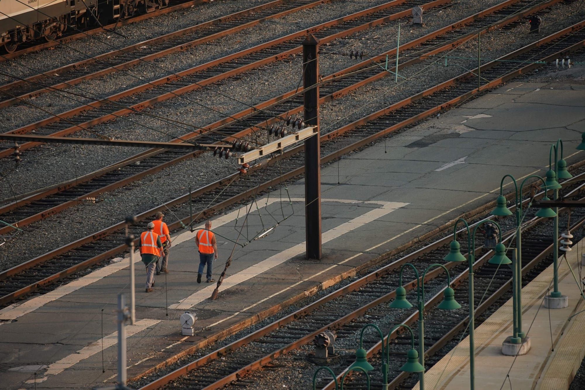 three railroad workers in orange vests and grey or blue pants walk between rail lines