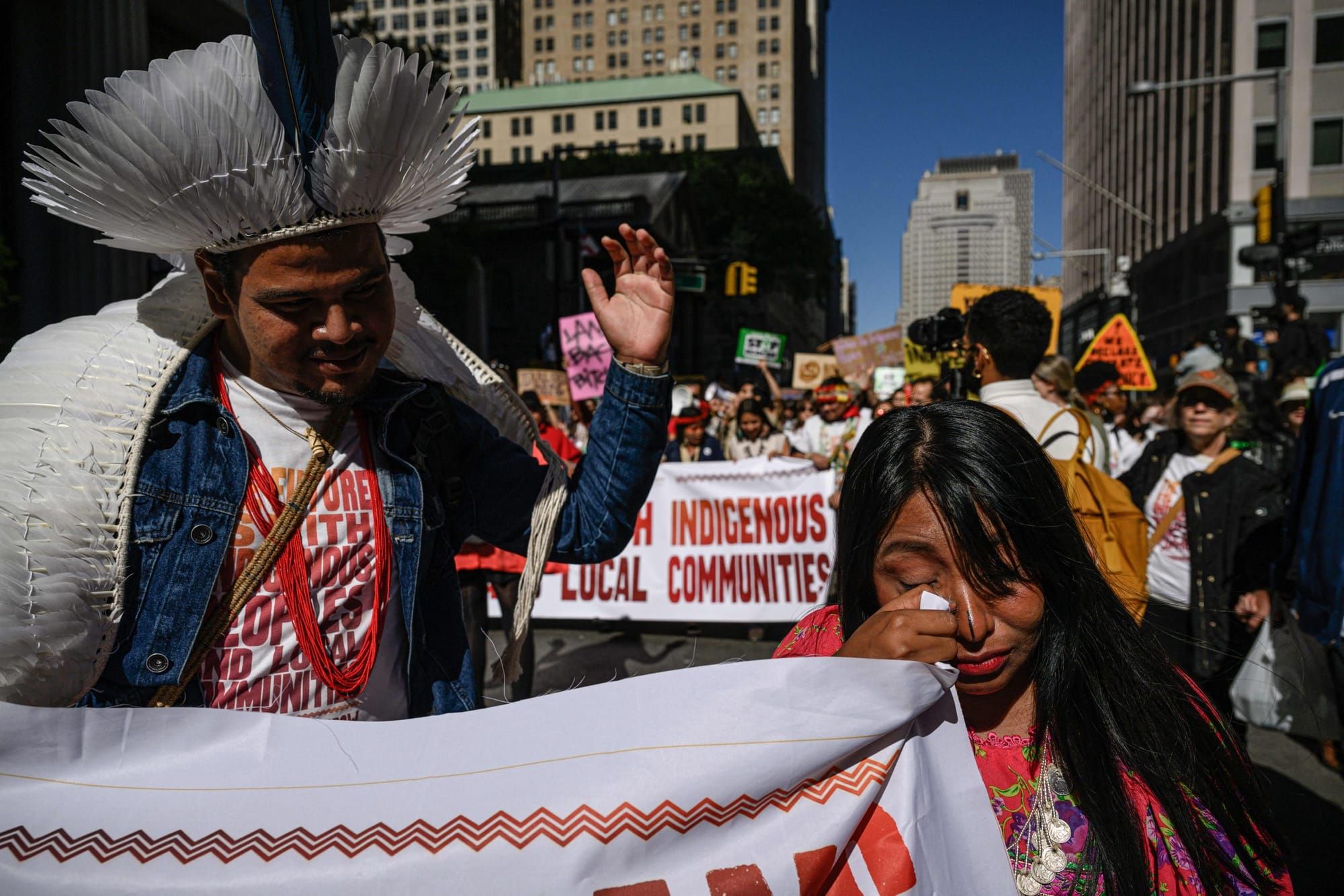 An Indigenous woman with bangs wipes tears from her eyes with the corner of a white cloth banner. Behind her, other activists