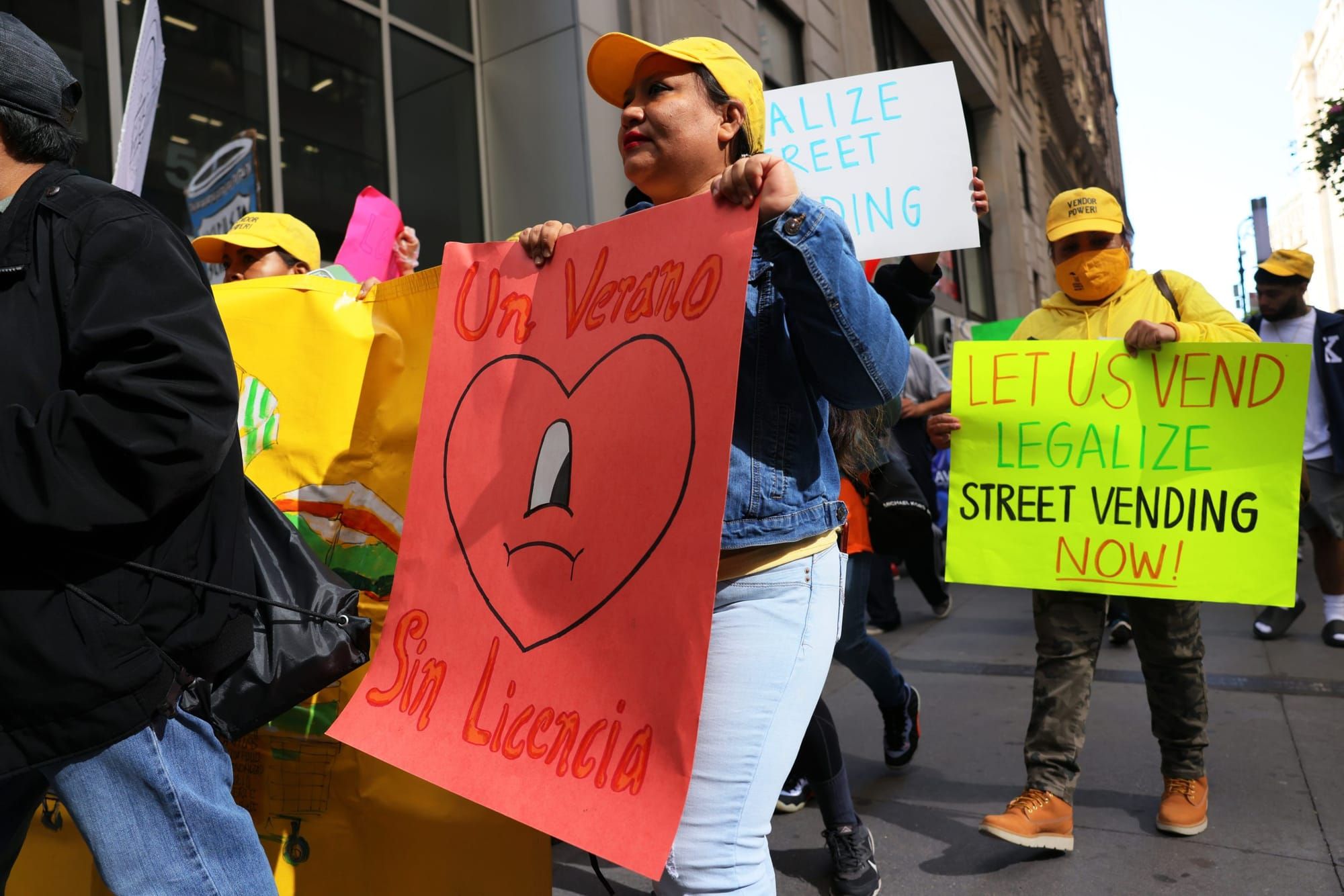 street vendors in yellow baseball caps hold large paper signs in different colors. one man in the background holds a yelllow