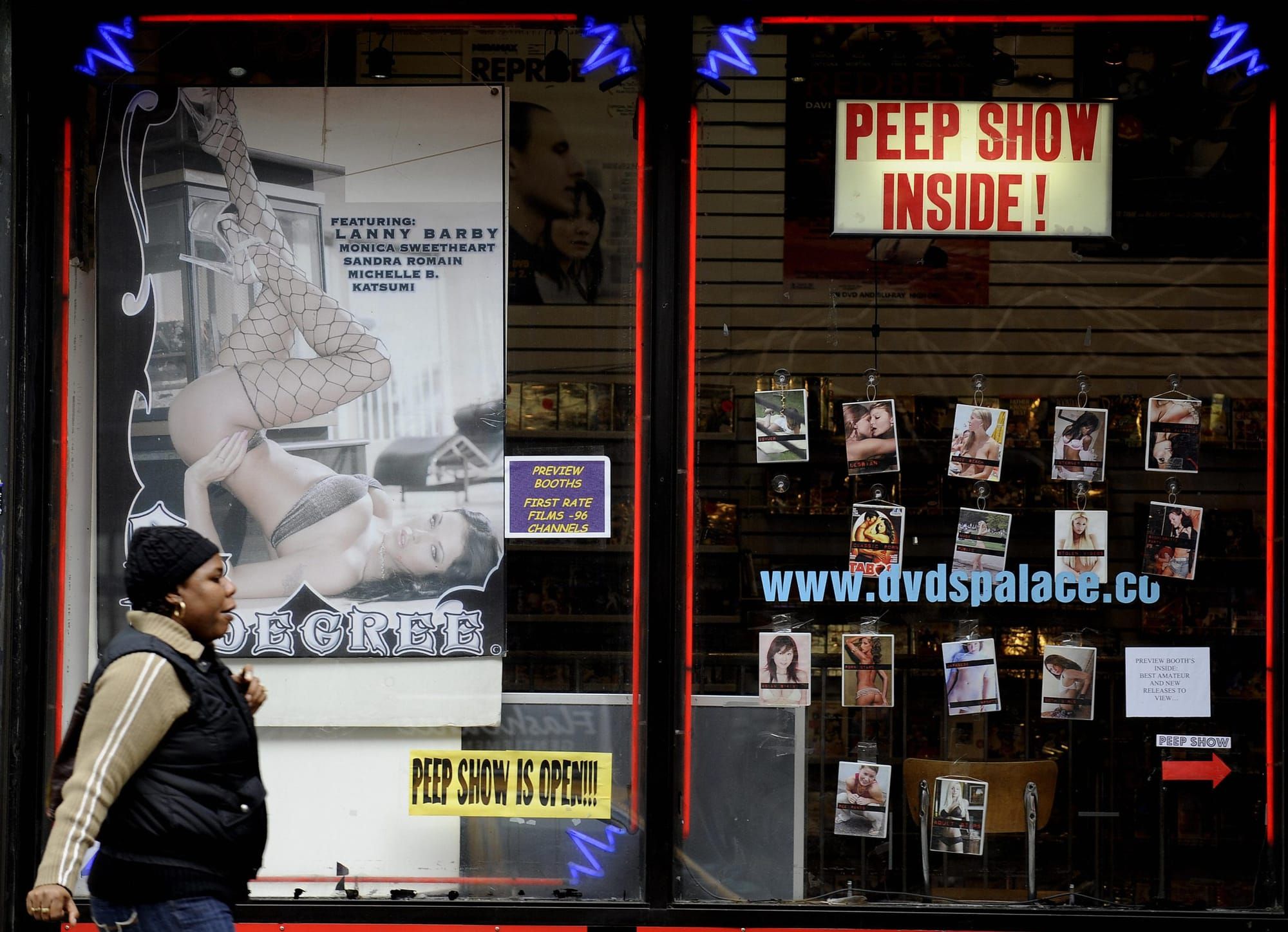 a Black woman in a light brown jacket and puffy black vest walks by the window of an adult video store. through the window a