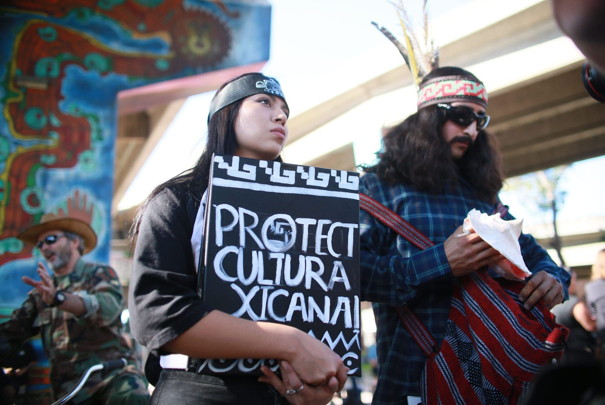 a Xicanx person with long black hair held back by a forehead band holds a black sign with white text that reads "protect cult