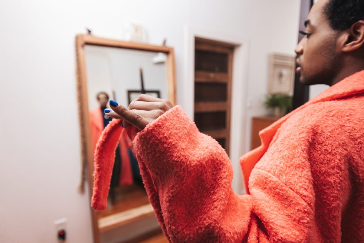 Home portrait of a young Black nonbinary person. They are wearing a pink colored jacket and looking at the mirror in the bedr