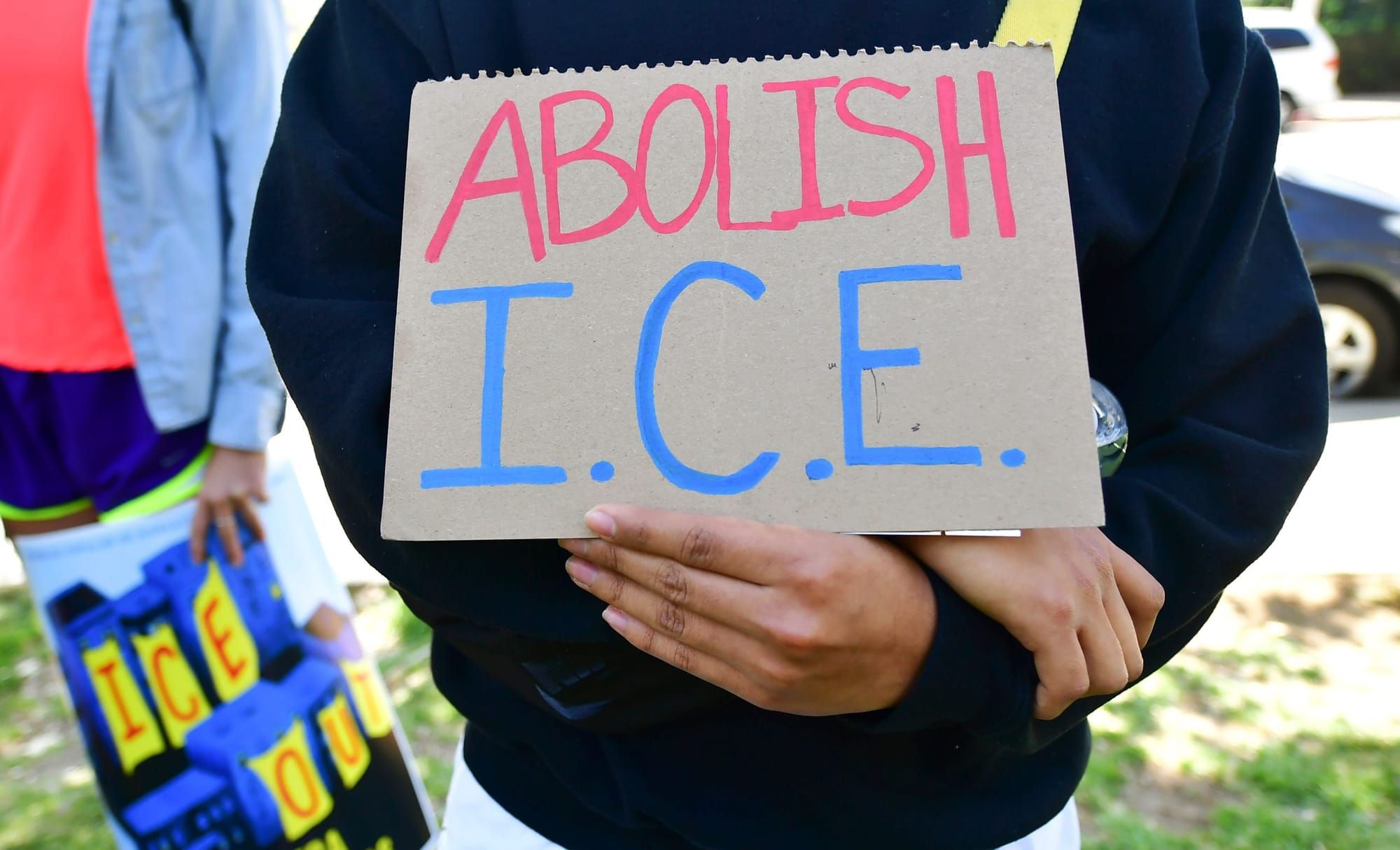 color photograph of a person wearing a black long-sleeve shirt holds a cardboard sign reading "Abolish ICE" in red and blue l